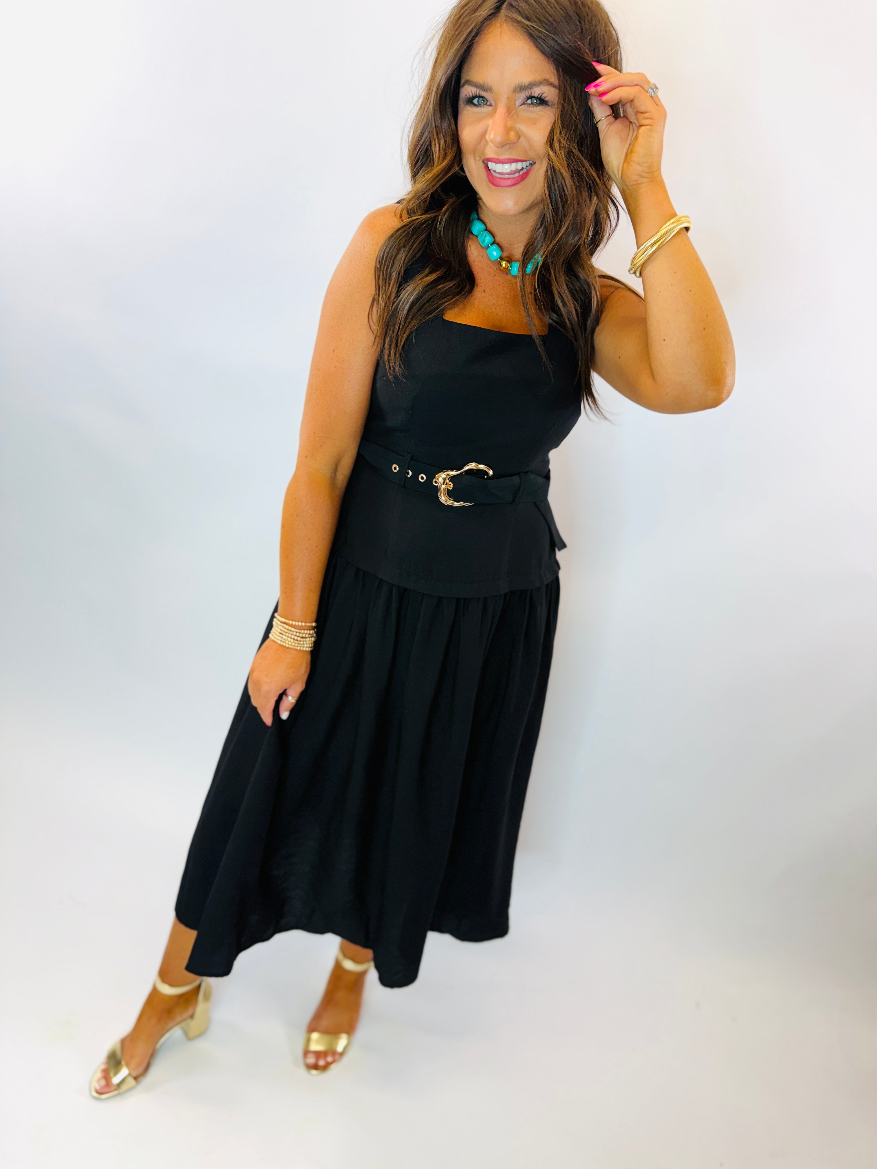 Woman wearing black sleeveless midi dress with gold belt buckle, paired with turquoise necklace and gold heels.