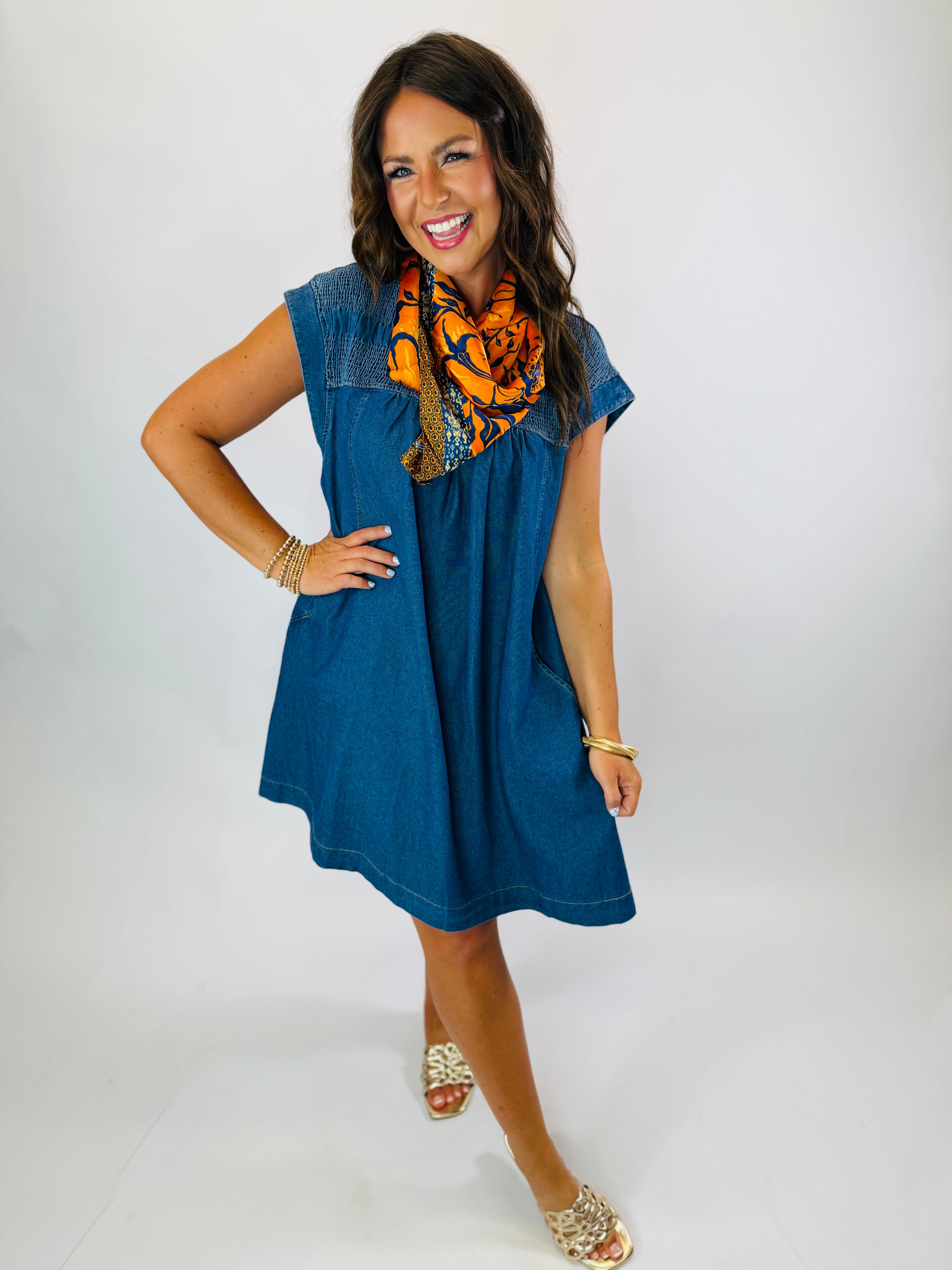 Woman wearing the Carson Dress, a dark denim short-sleeve babydoll dress with smocked shoulders, styled with an orange flo...
