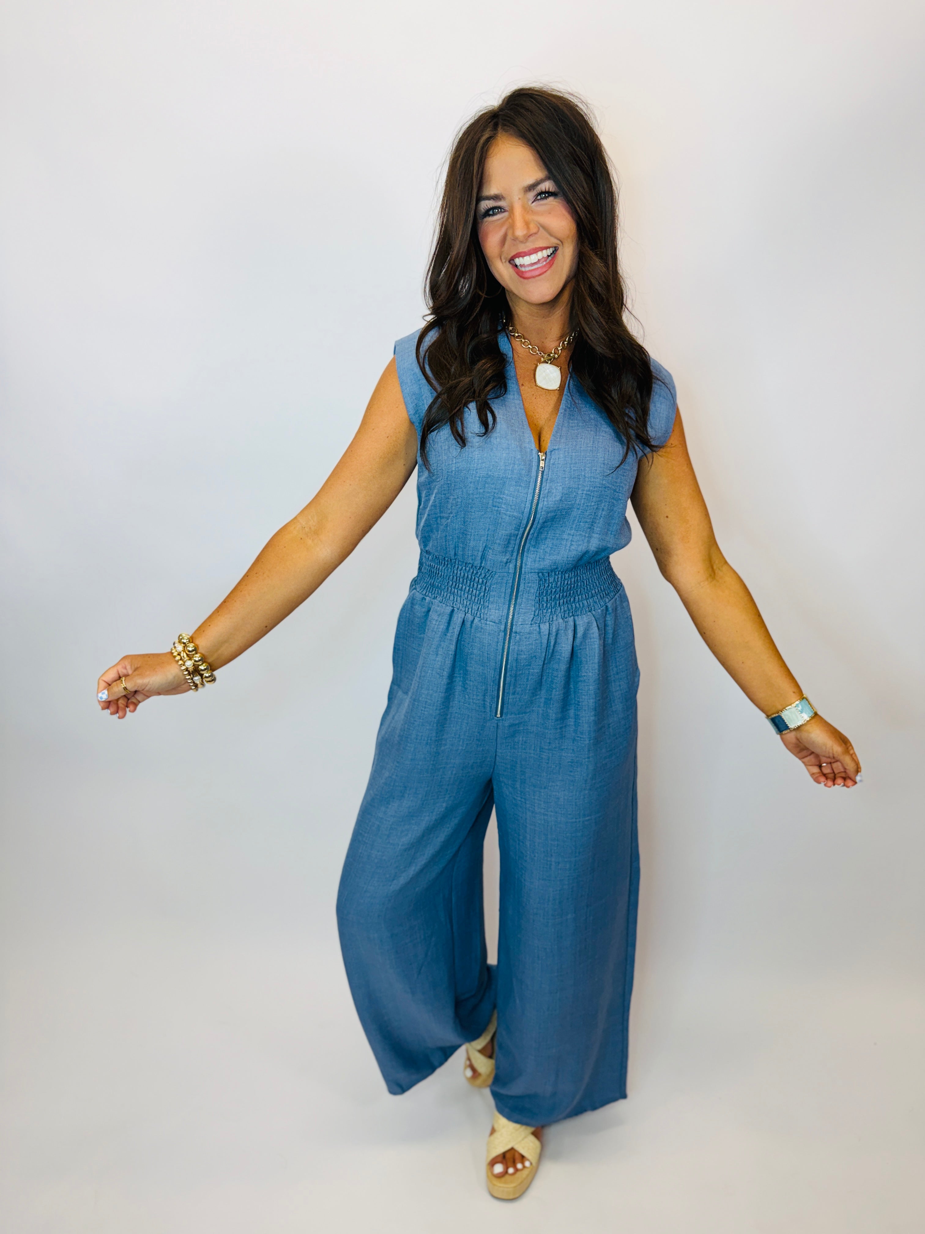 Woman wearing blue denim sleeveless jumpsuit with zip front and wide-leg pants, styled with gold jewelry.