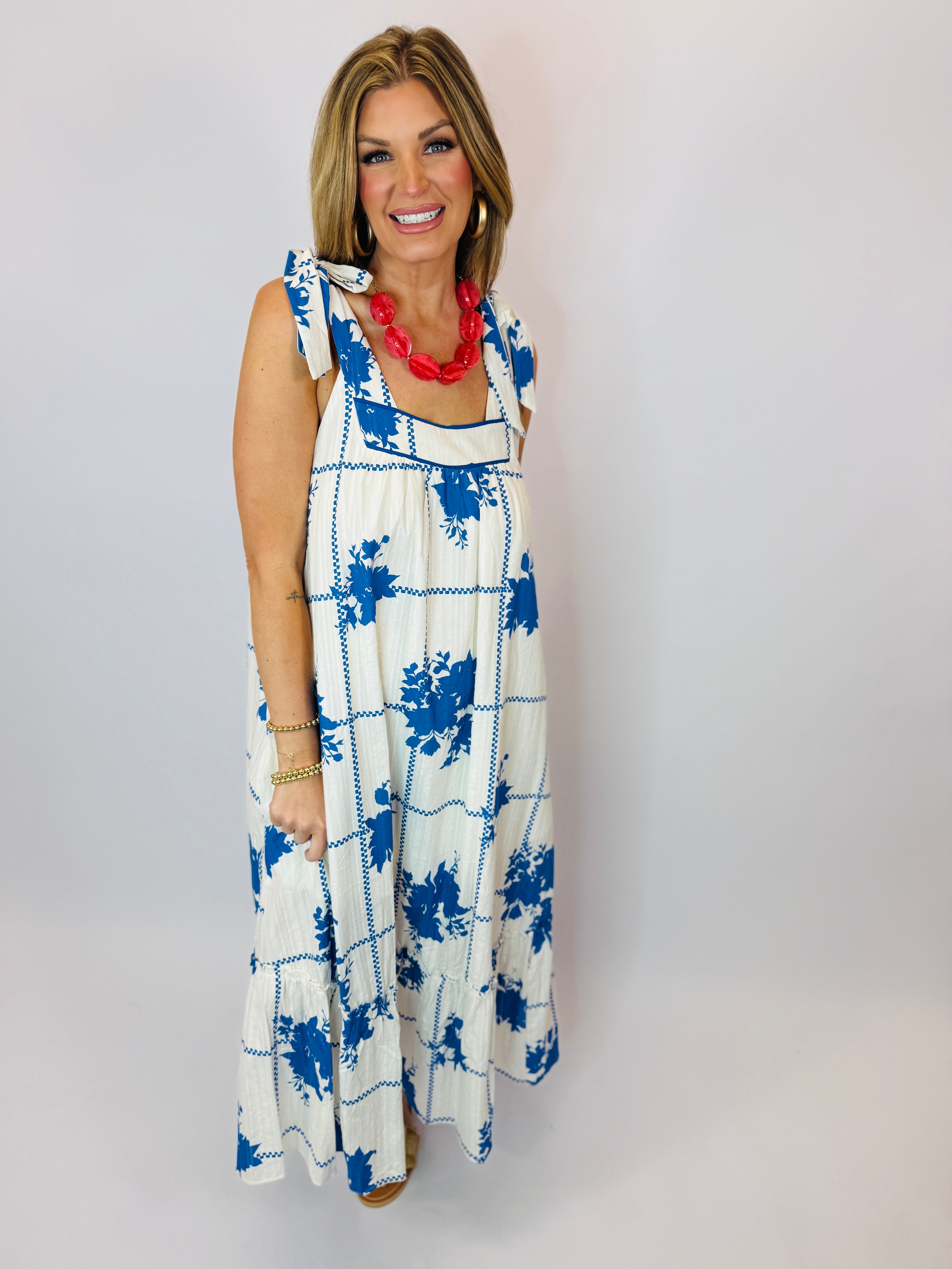 White maxi dress with blue floral print, tie shoulder straps, and tiered design modeled by smiling woman