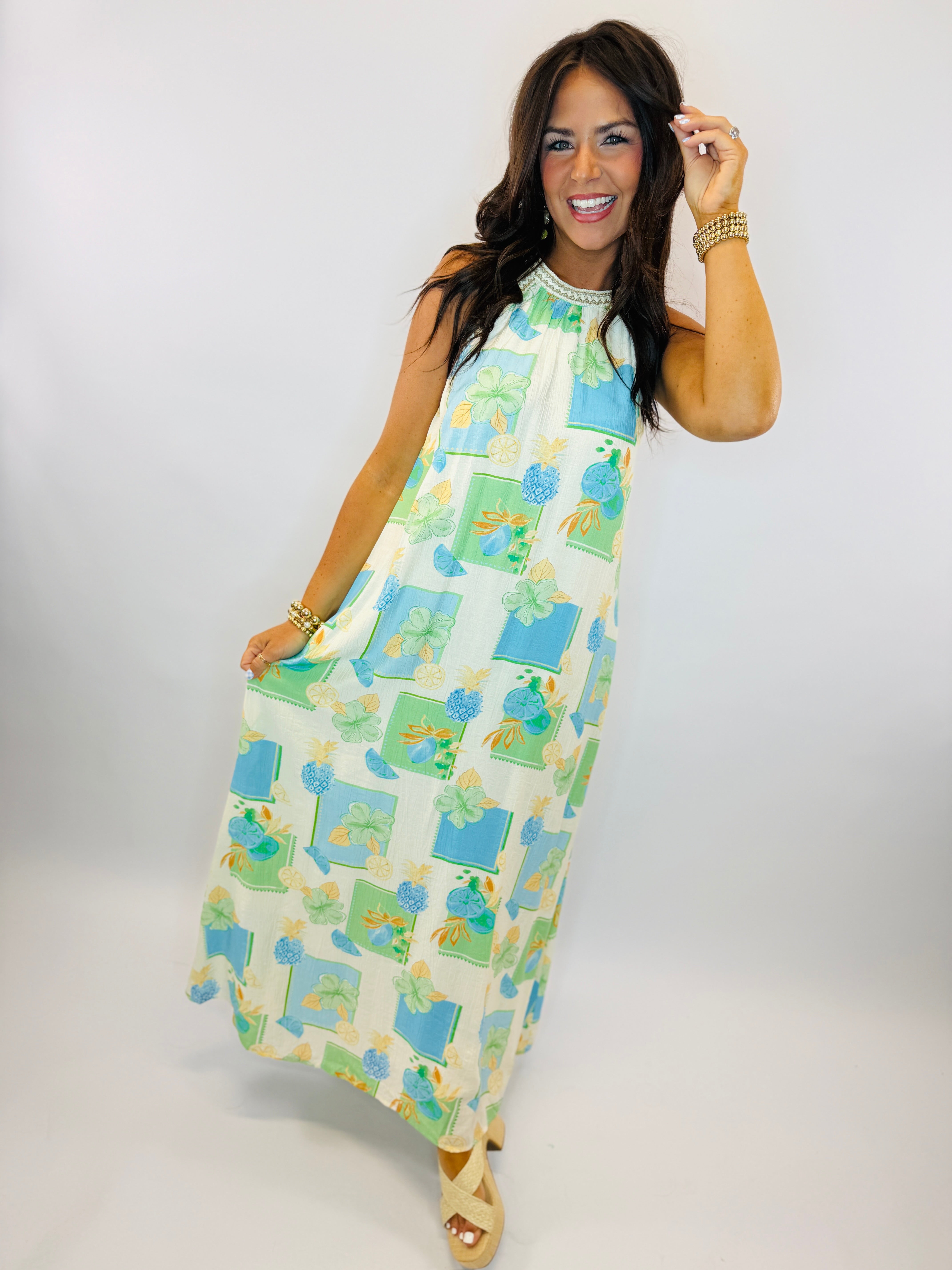 Woman wearing colorful sleeveless Bermuda dress with blue and green floral pattern, styled with gold jewelry and wedge san...