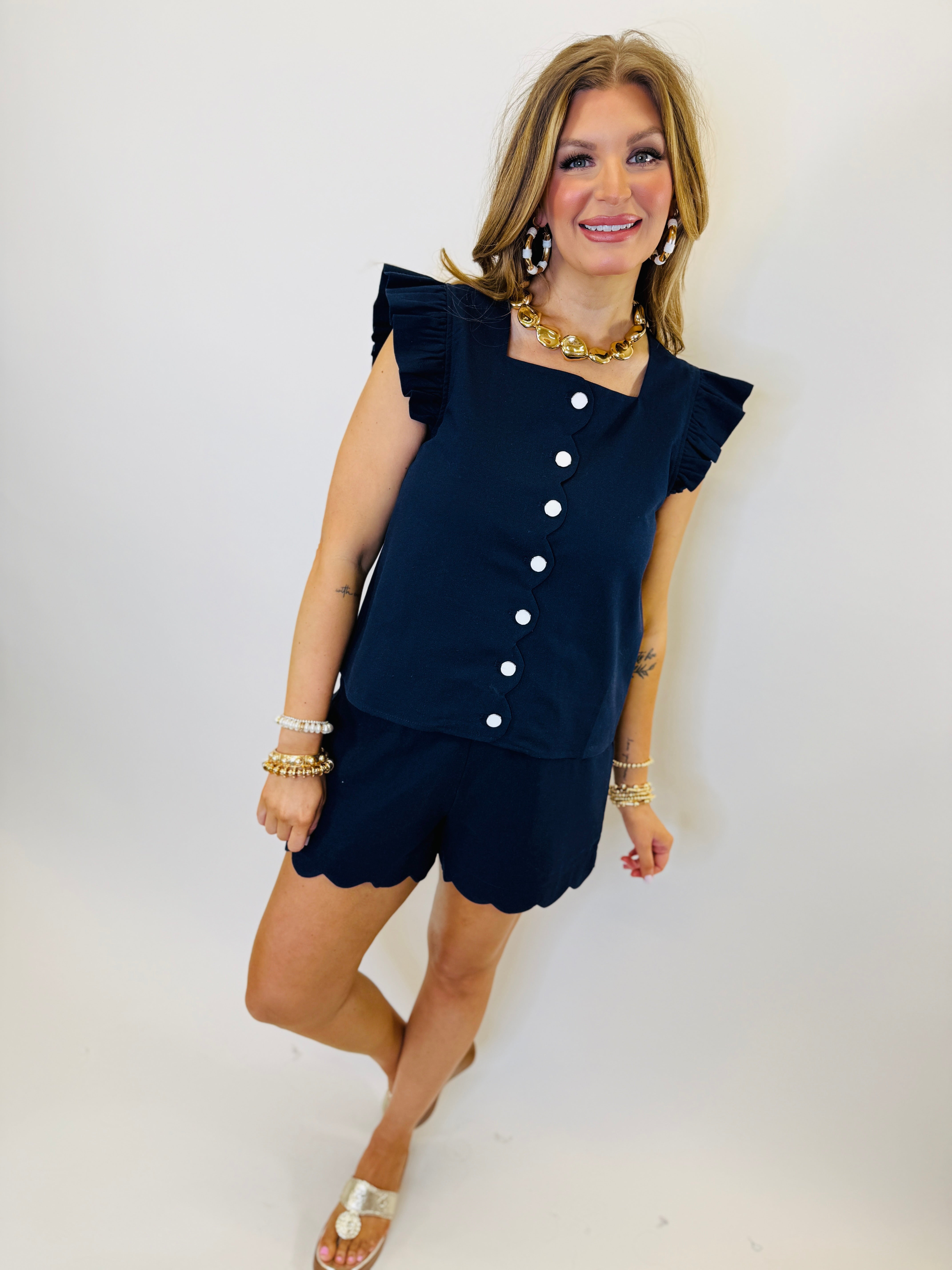 Navy blue ruffle sleeve top with white buttons and scalloped hem, styled with matching shorts and gold jewelry.