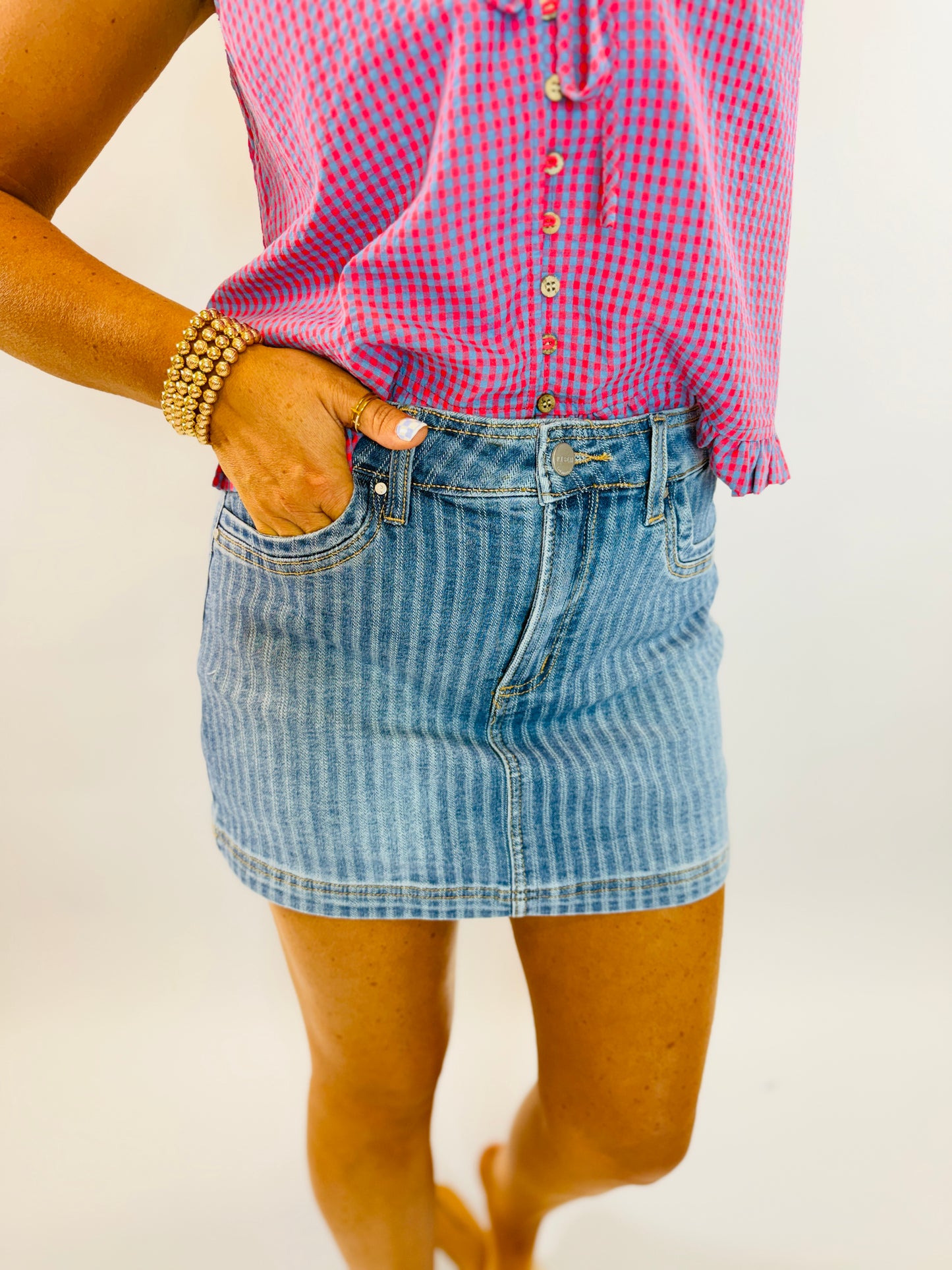 Woman wearing striped denim mini skort with hand in pocket, paired with pink gingham shirt and gold bracelet.