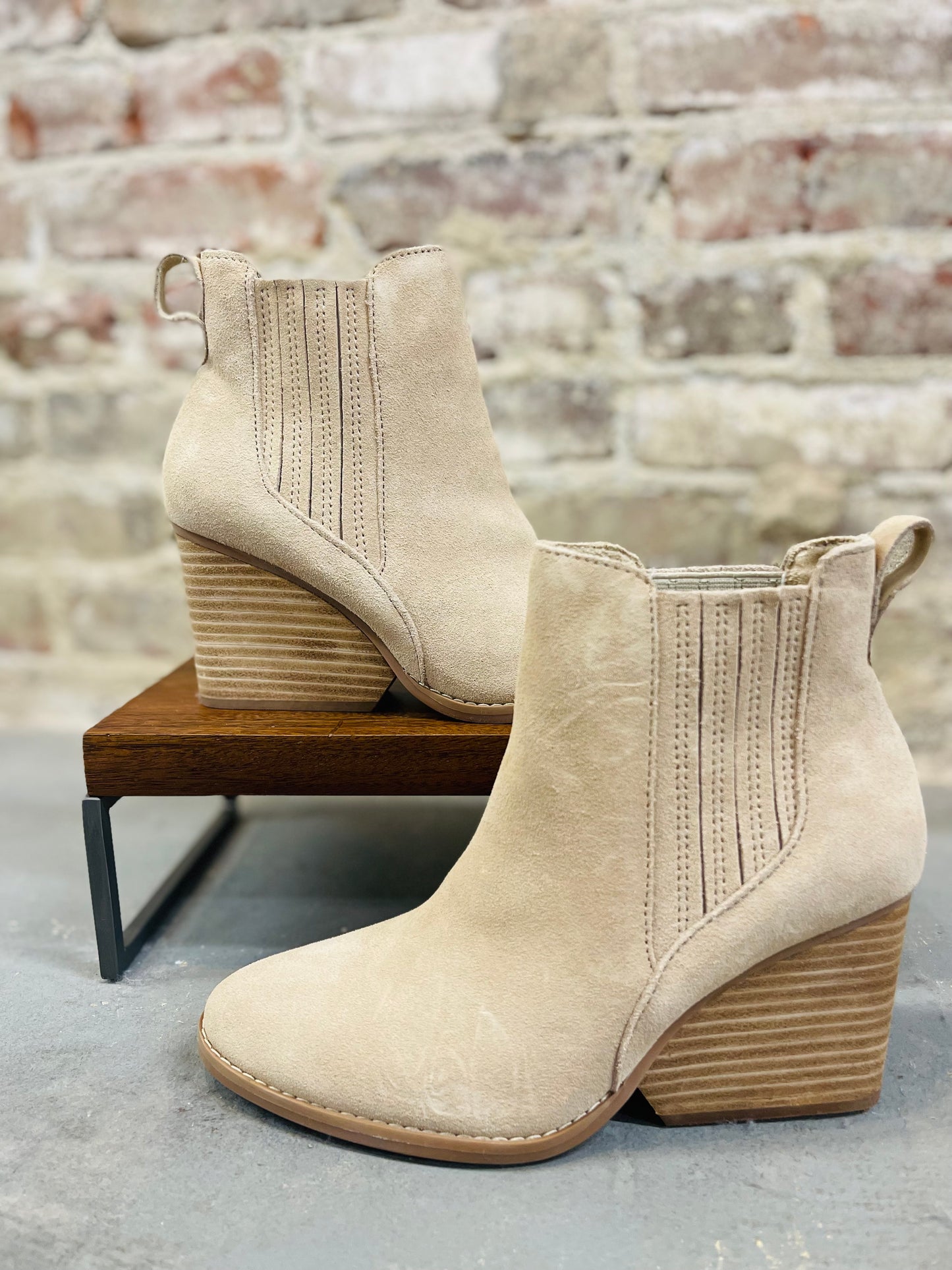 Beige suede Noa ankle boots with stacked wooden heels and elastic side panels displayed on wooden bench.