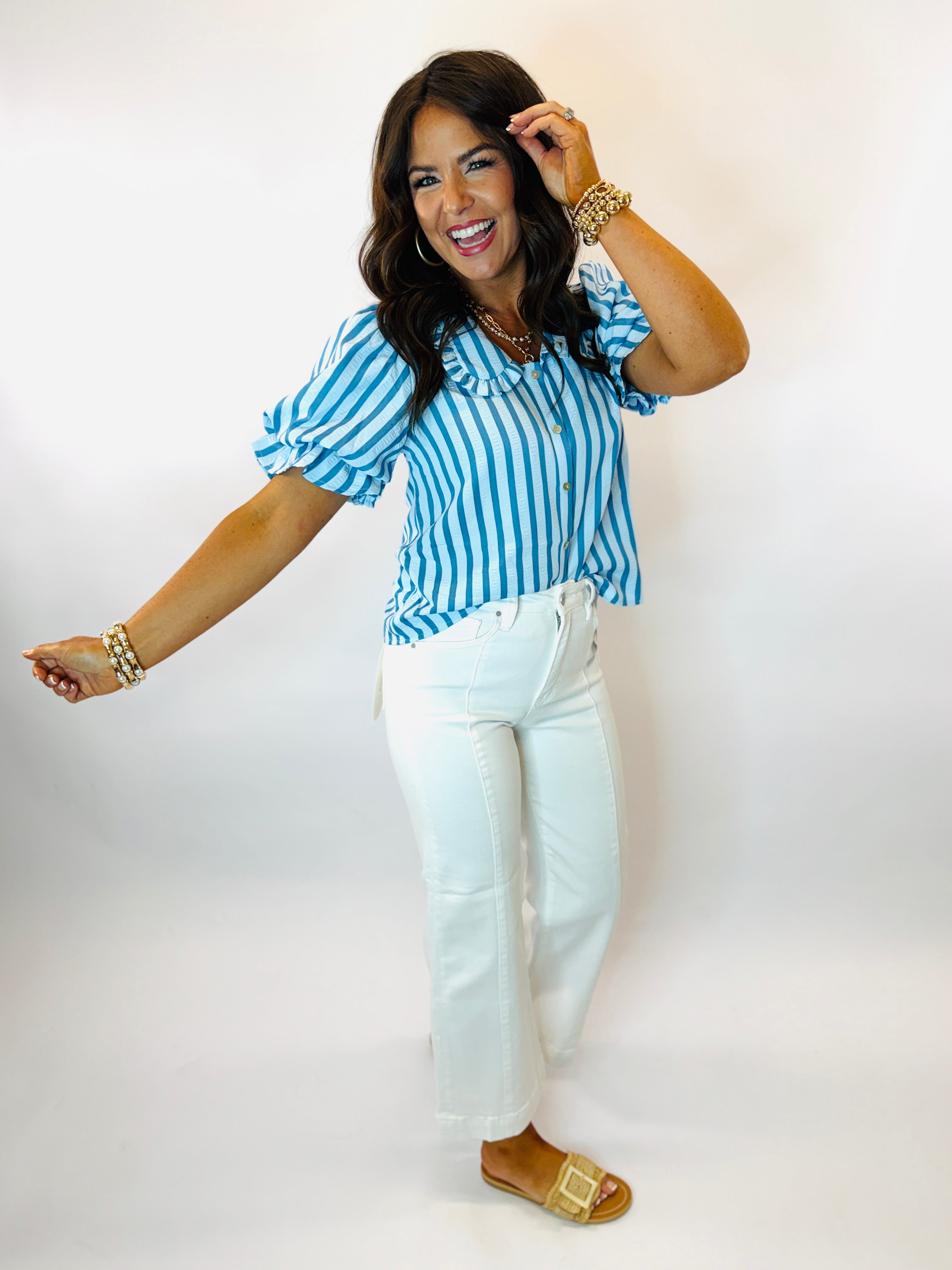 Woman wearing blue and white striped button-up blouse with puffy sleeves, white jeans, and gold accessories