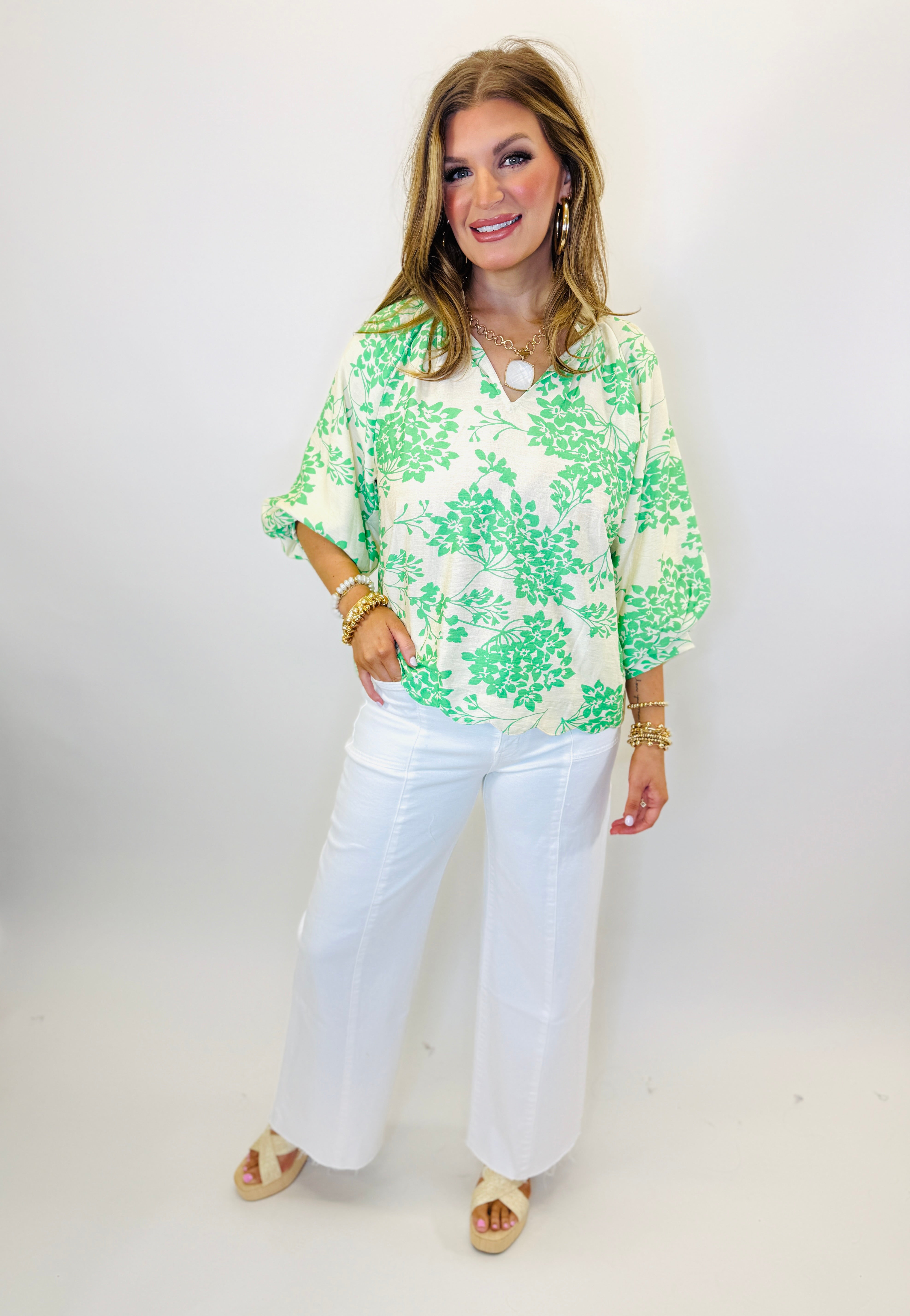 Woman wearing green and white floral print Amelia top with white wide-leg pants and gold jewelry.