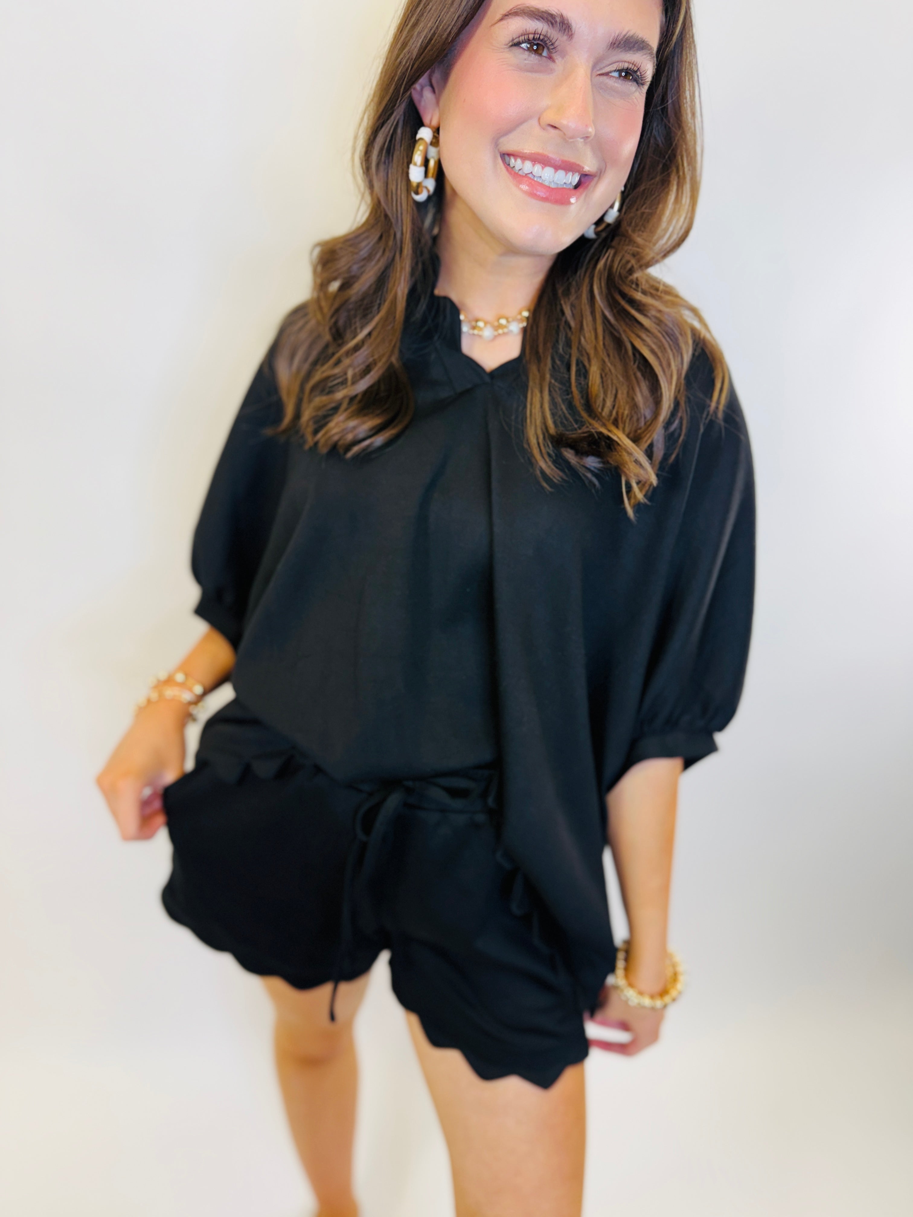 Woman wearing black oversized collared top with tie waist and matching shorts, styled with pearl jewelry.