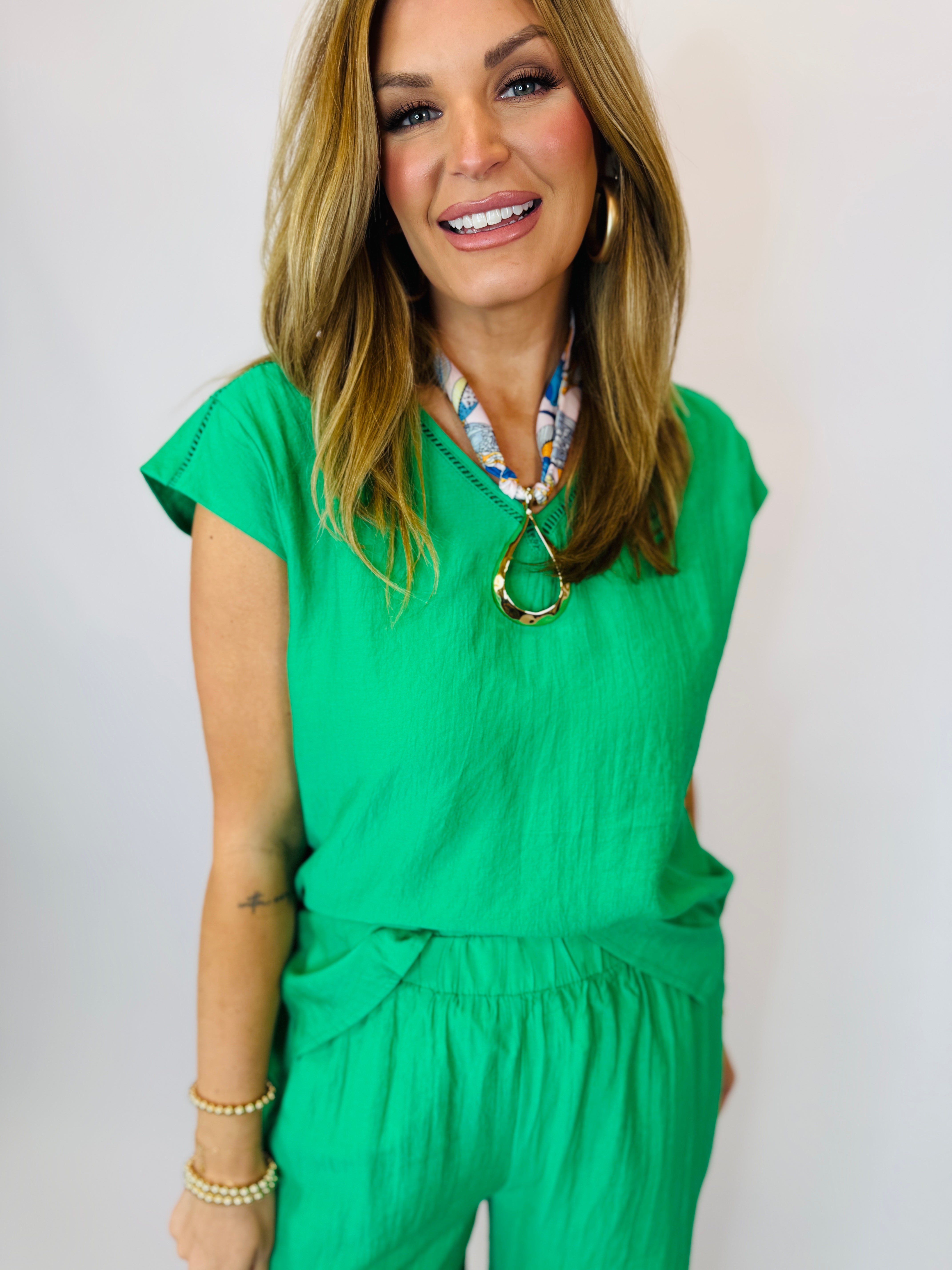 Woman wearing bright green short-sleeve top with v-neck and tie waist detail, styled with layered necklaces.