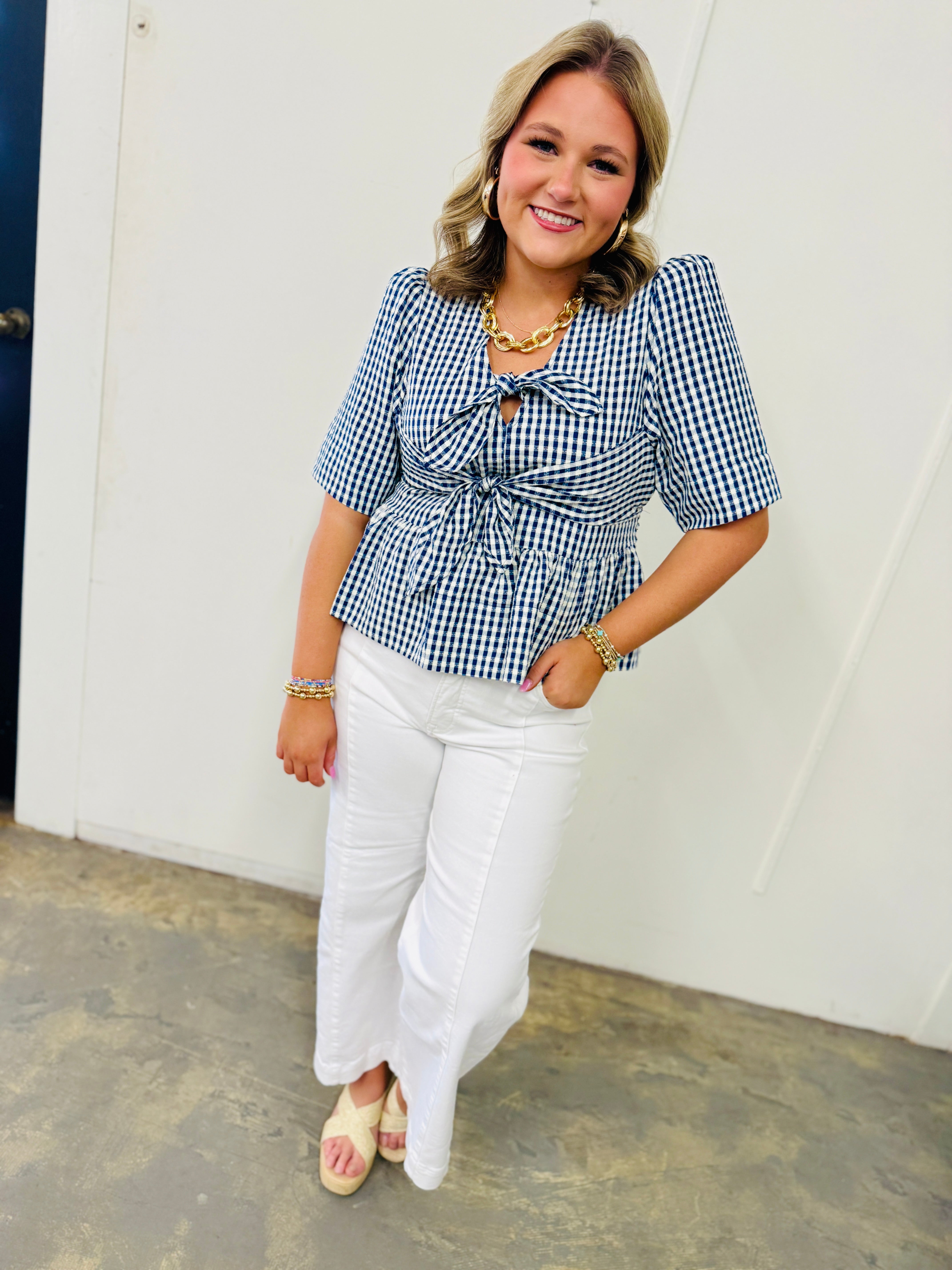 Woman wearing blue and white gingham tie-front top with white jeans and gold chain necklace