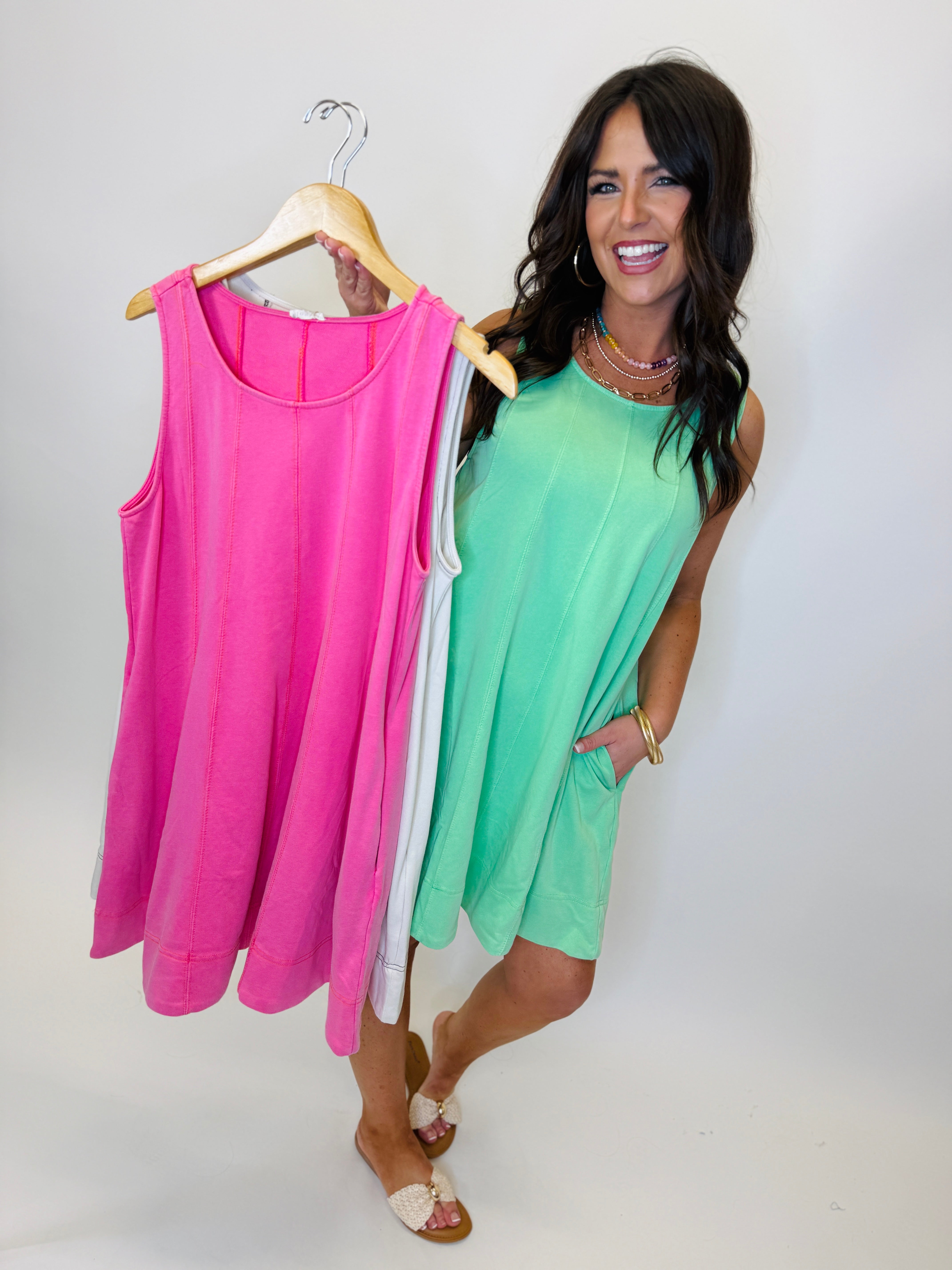 Woman modeling bright green sleeveless dress while holding pink version on hanger, showing both color options available.