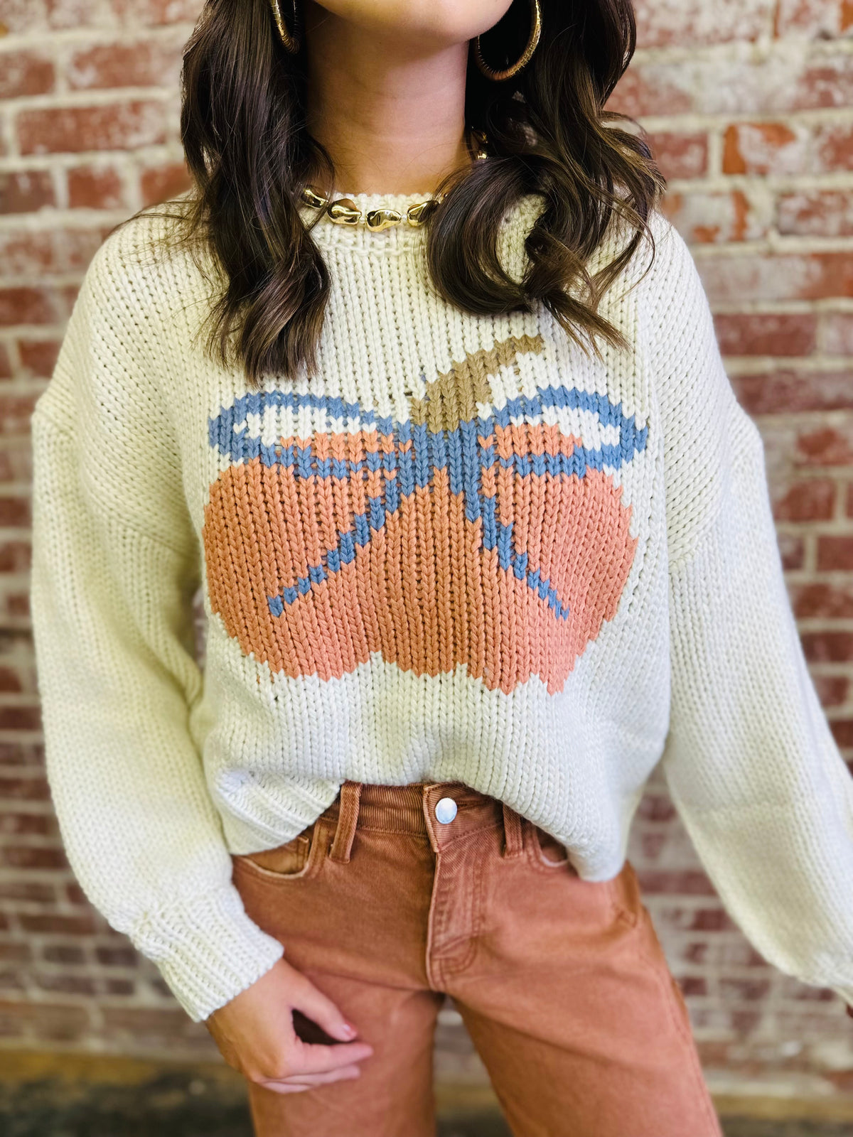 Cream knit sweater with colorful pumpkin design, worn with brown corduroy pants against brick wall background.
