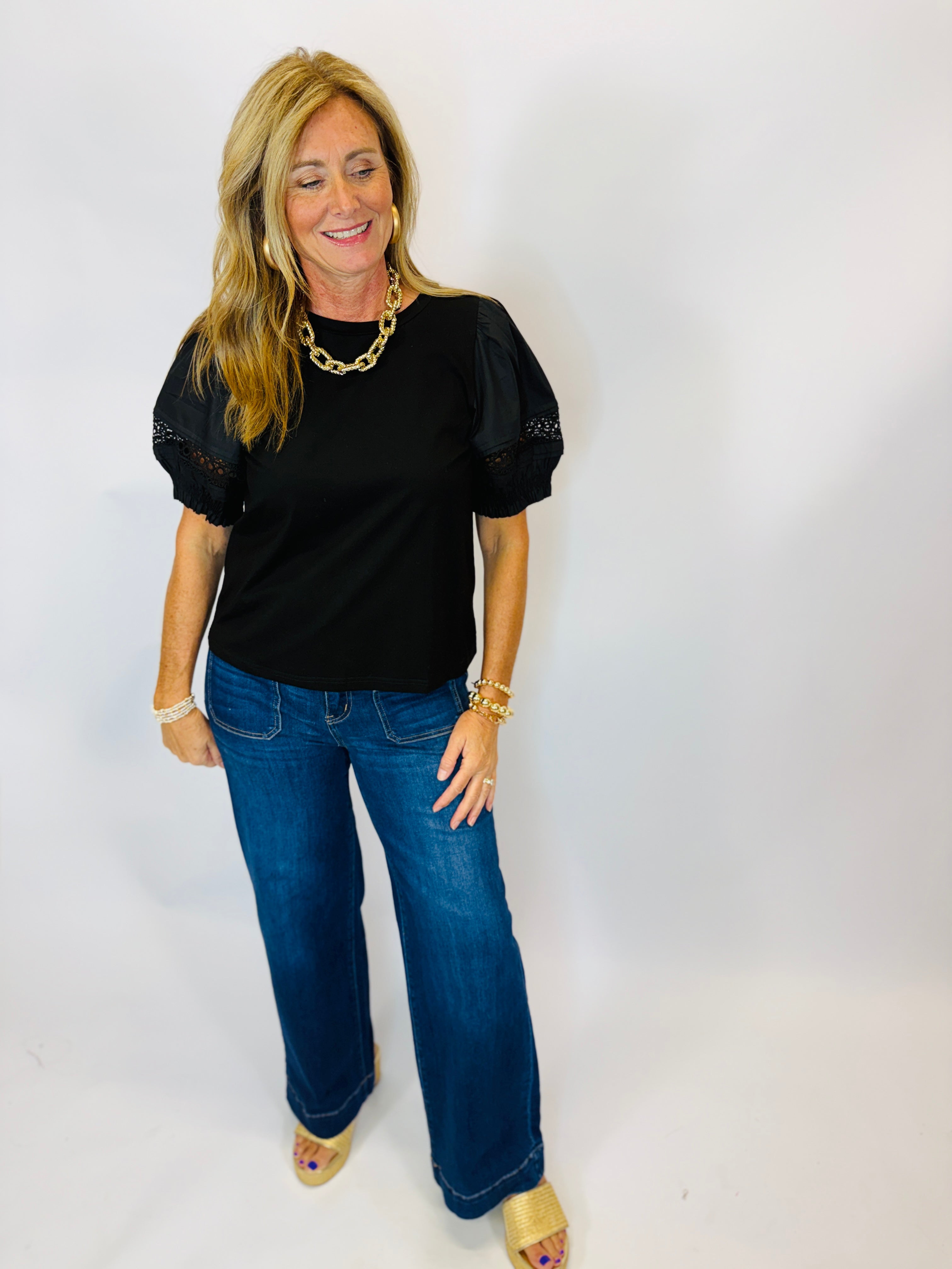 Black short-sleeve Jenna Top with lace puff sleeves, styled with wide-leg jeans and gold accessories.