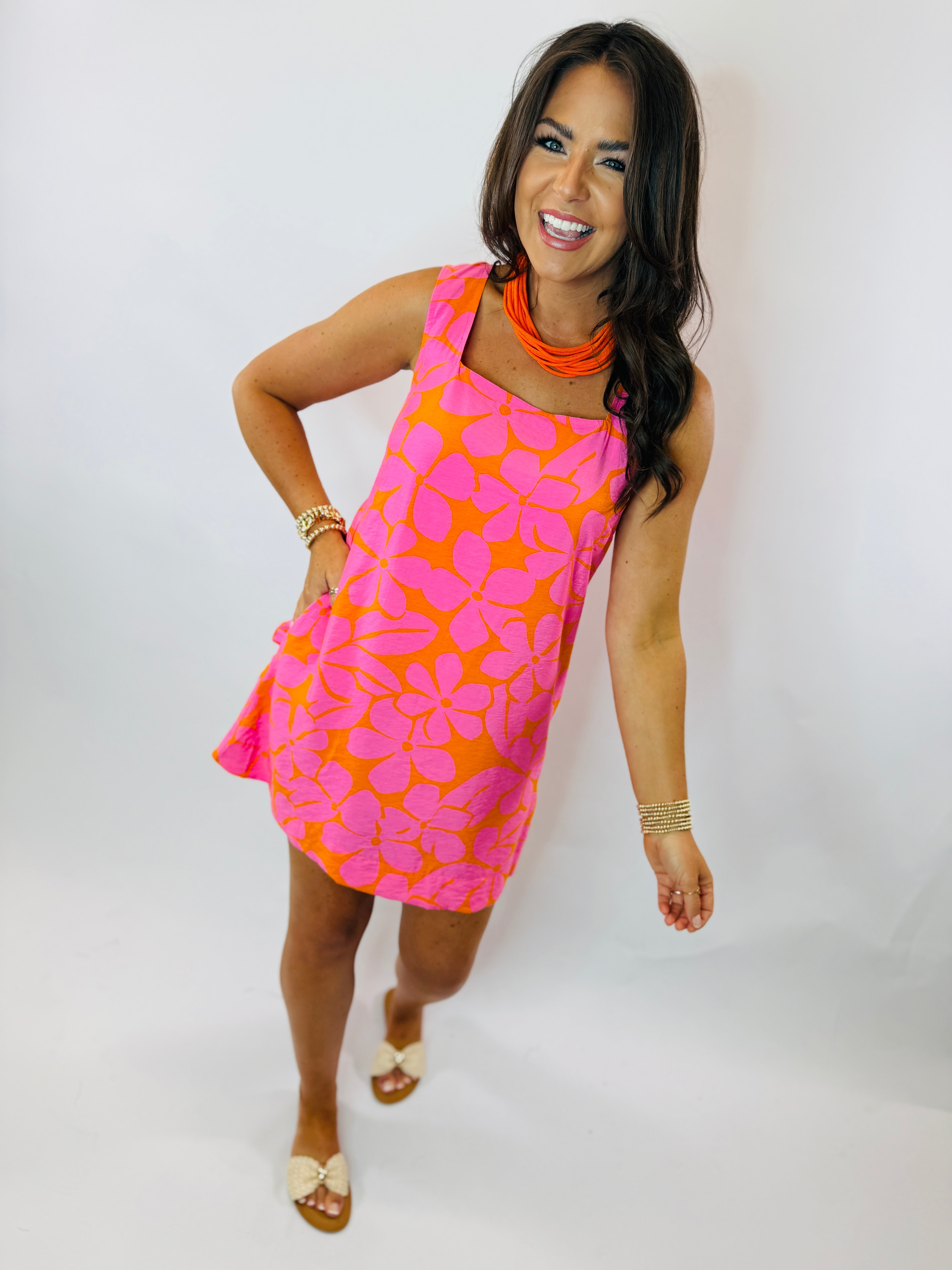 Woman wearing bright pink and orange floral patterned sleeveless mini dress with orange statement necklace and gold bracel...