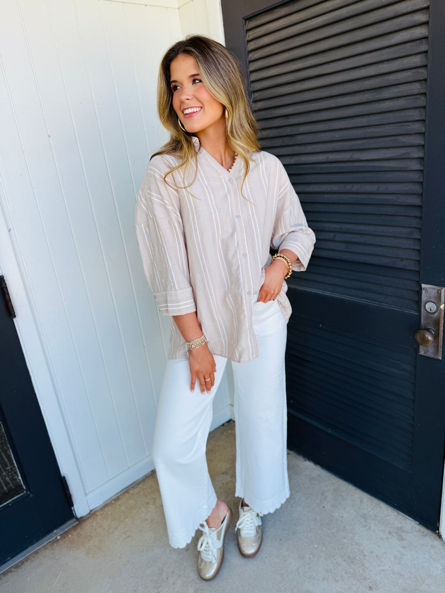 Woman wearing beige striped button-up shirt with white wide-leg pants and gold jewelry, smiling outdoors