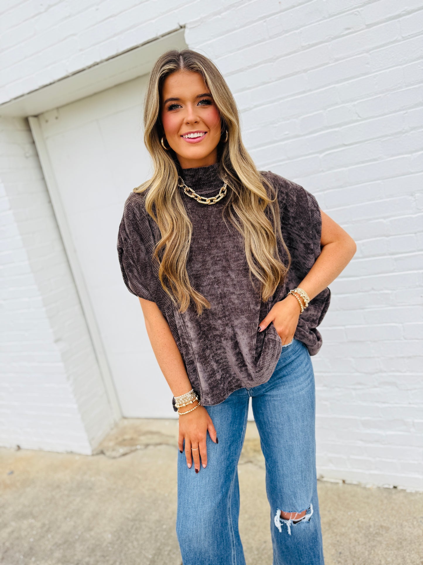 Woman wearing charcoal gray oversized knit turtleneck top with short sleeves, paired with distressed jeans and gold jewelry.