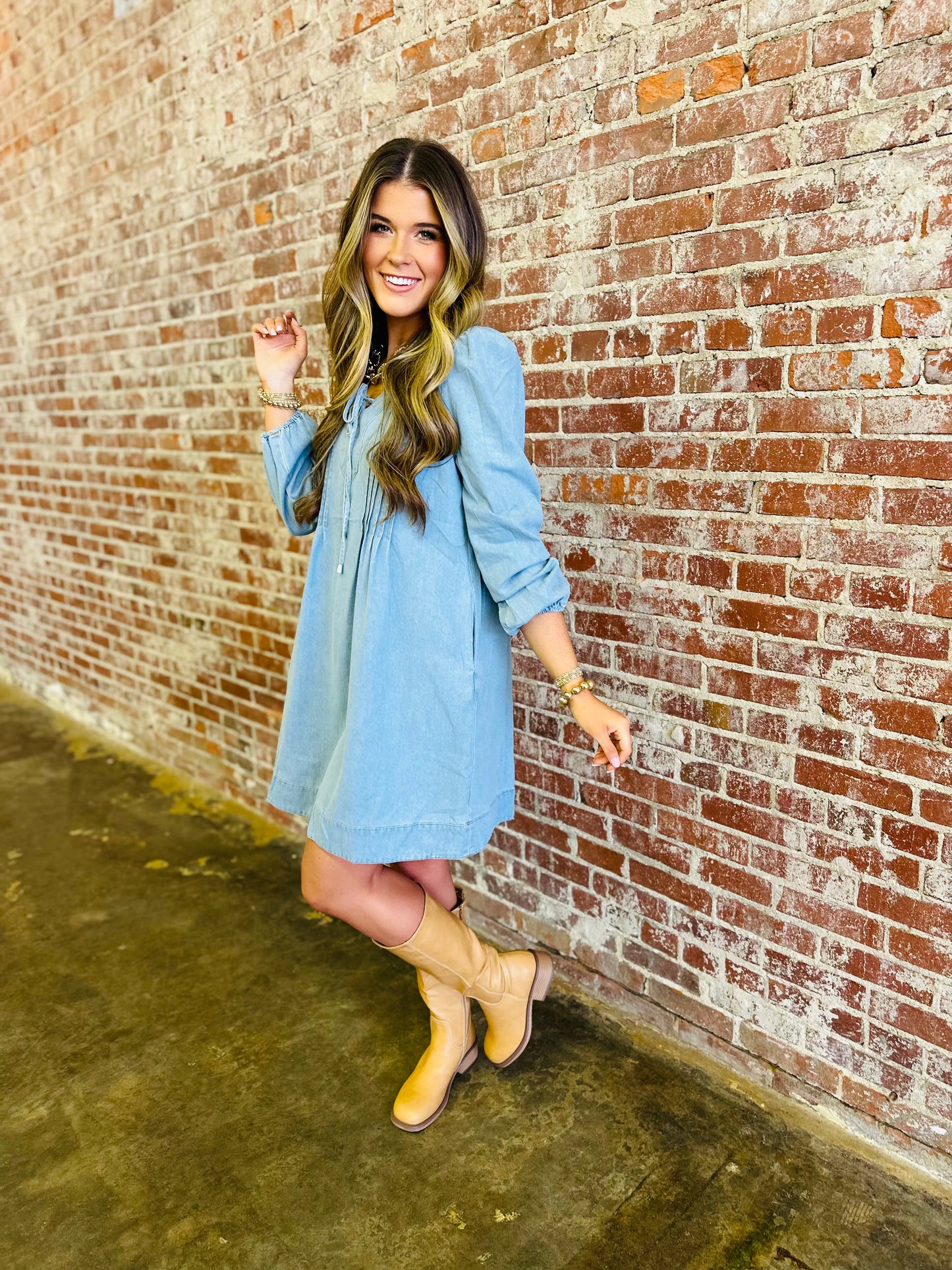 Woman wearing light blue denim dress with long sleeves and drawstring neckline, paired with tan cowboy boots.