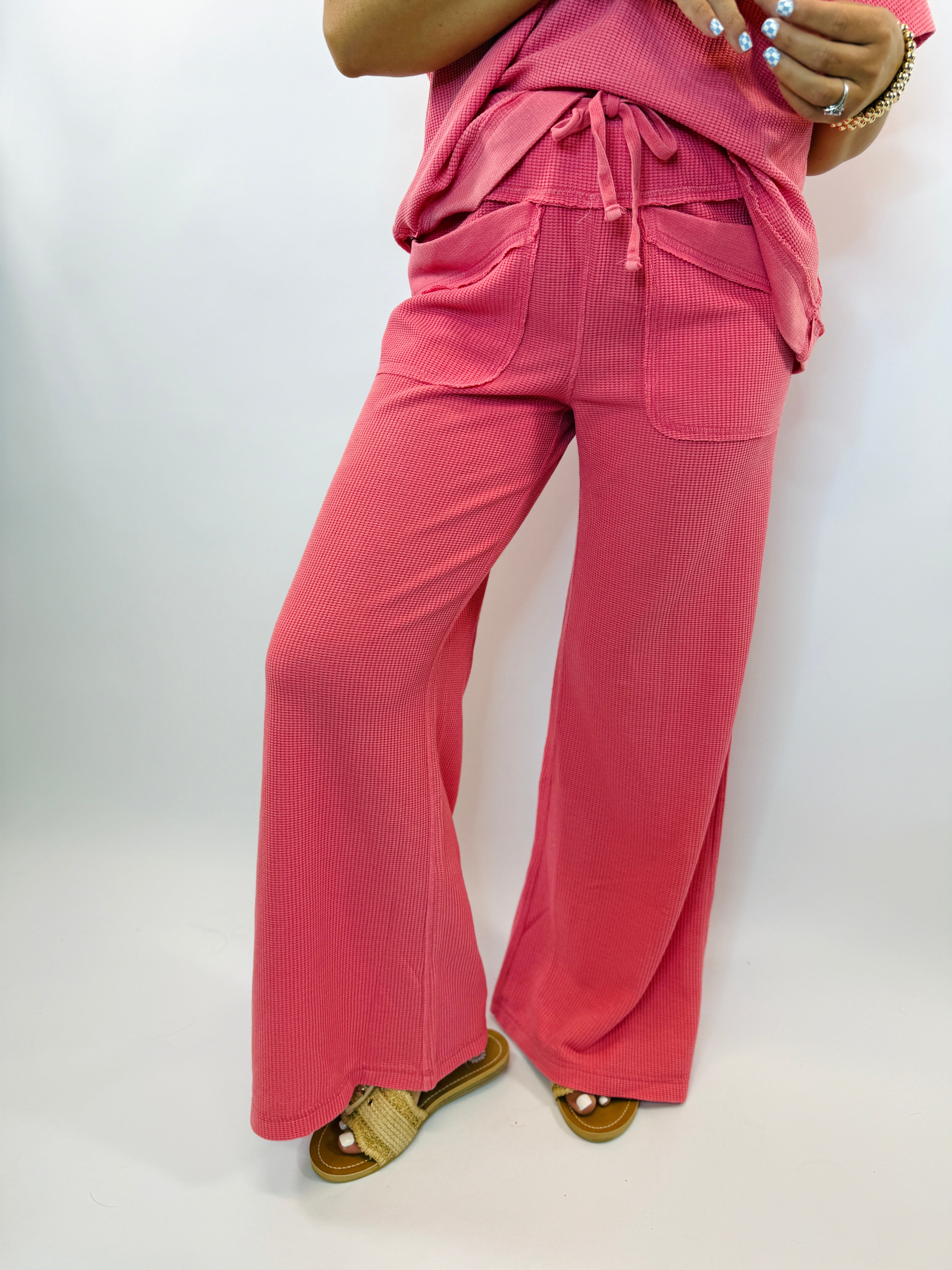 Pink ribbed wide-leg pants with drawstring waist and cargo pockets, paired with gold woven sandals.