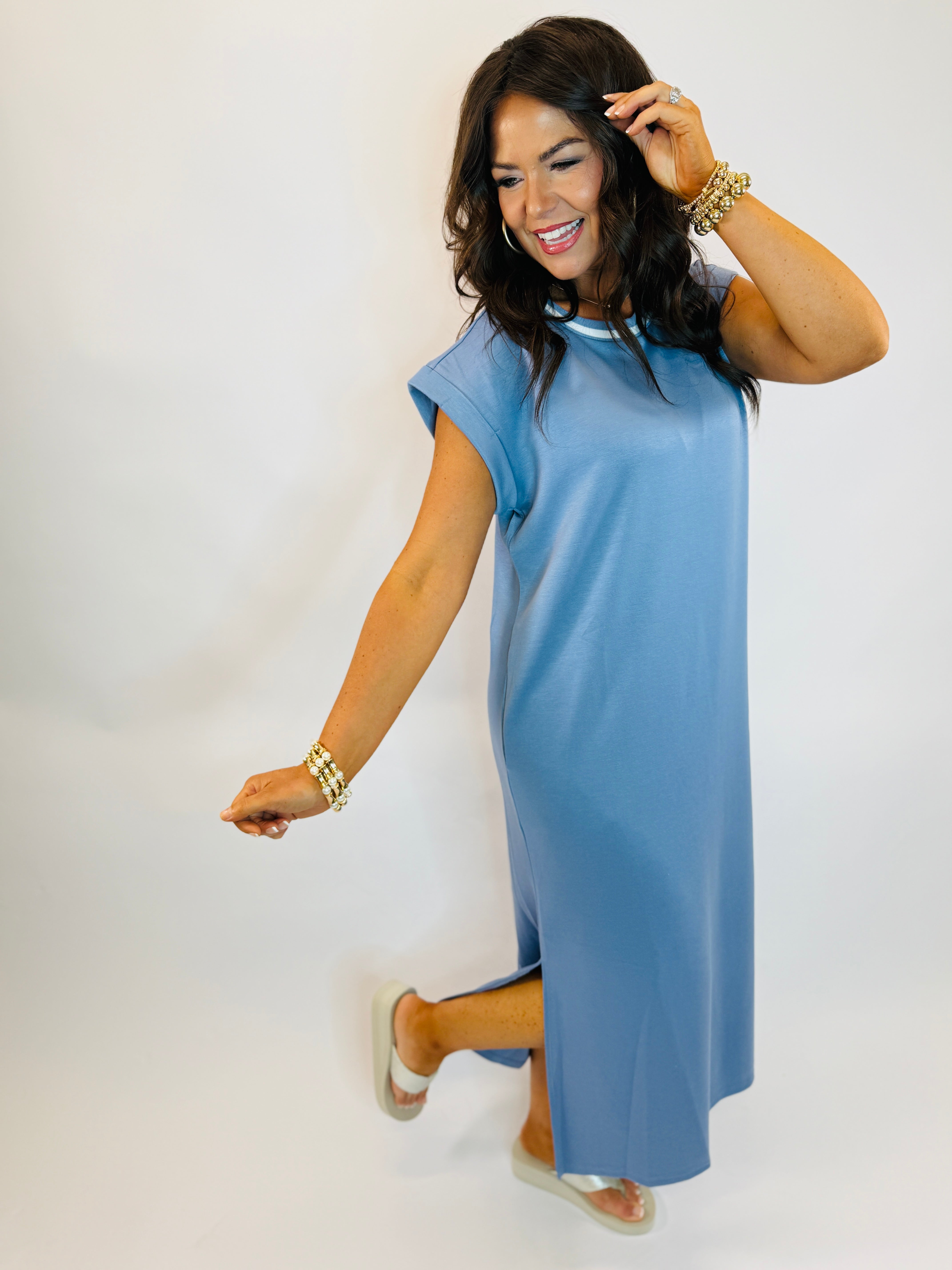 Woman wearing blue short-sleeve midi dress with gold jewelry and sandals, smiling and posing against white background.