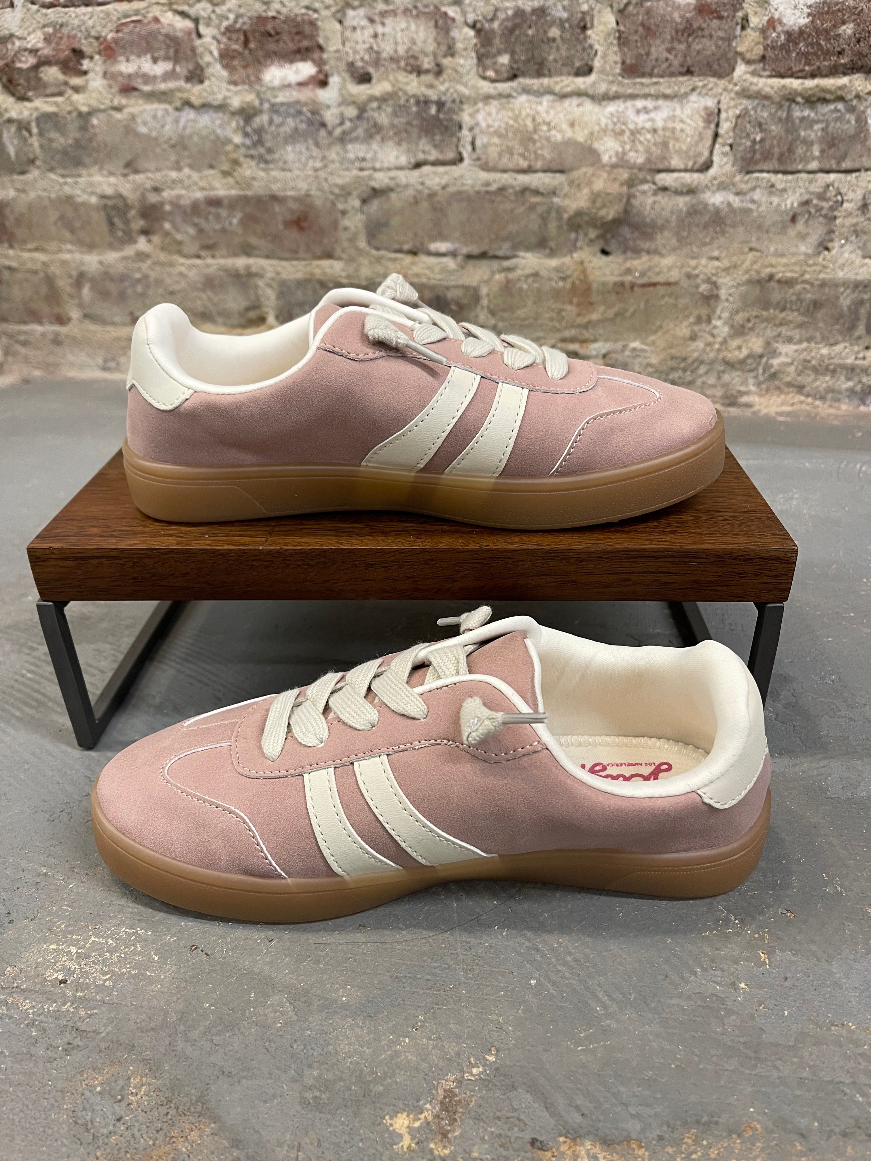 Pink suede sneakers with cream accents and gum sole displayed on wooden platform against brick wall