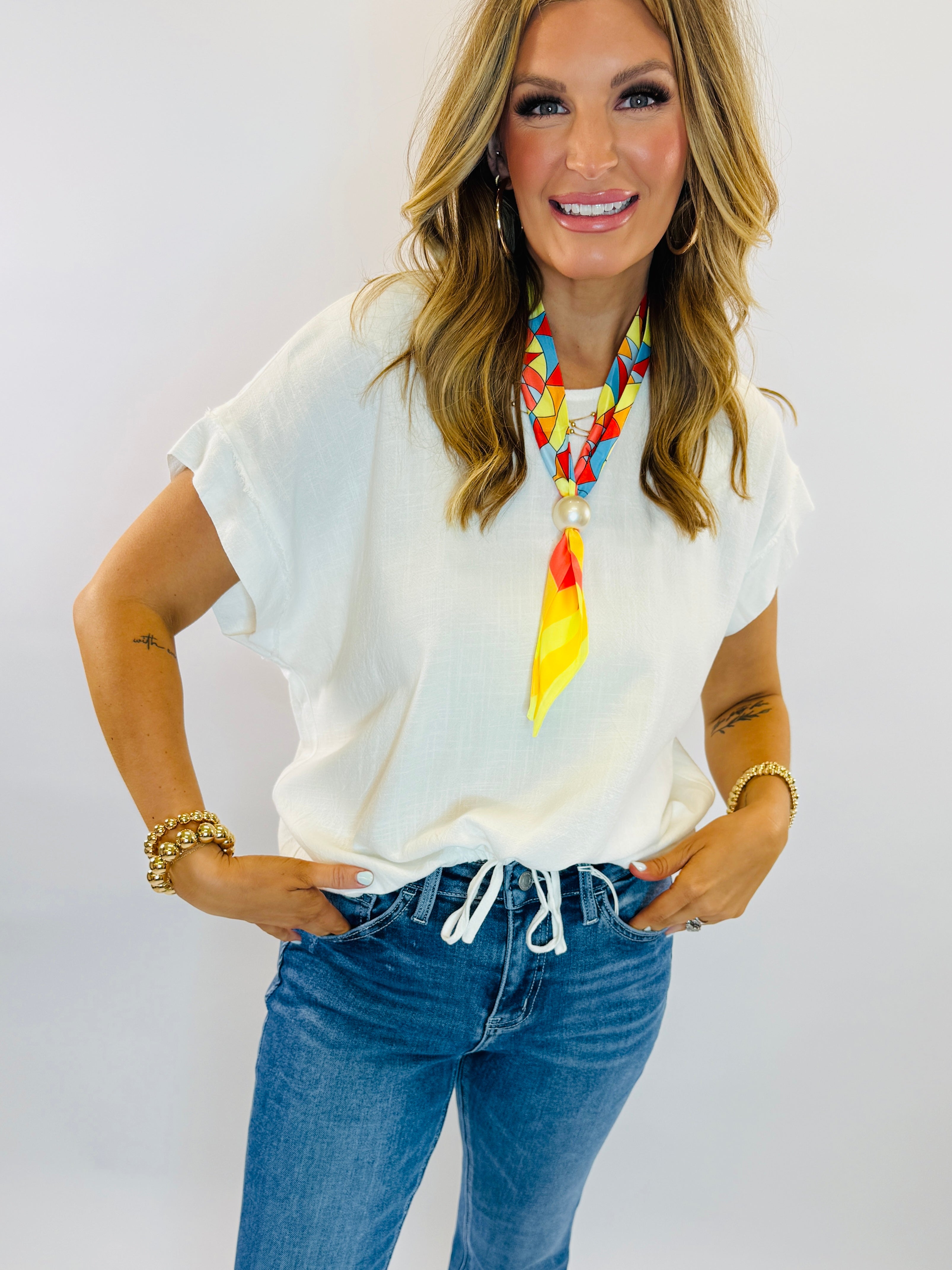 Woman wearing white linen short-sleeve top with drawstring hem, styled with blue jeans and colorful statement necklace