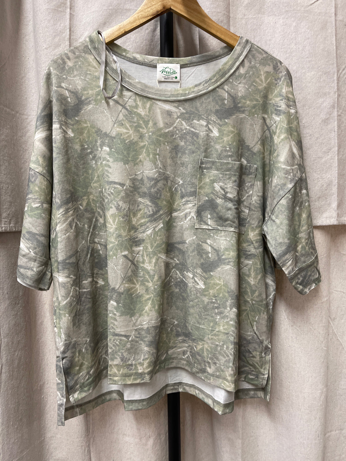 Sage green tie-dye long sleeve top with chest pocket hanging on wooden hanger against curtain backdrop.