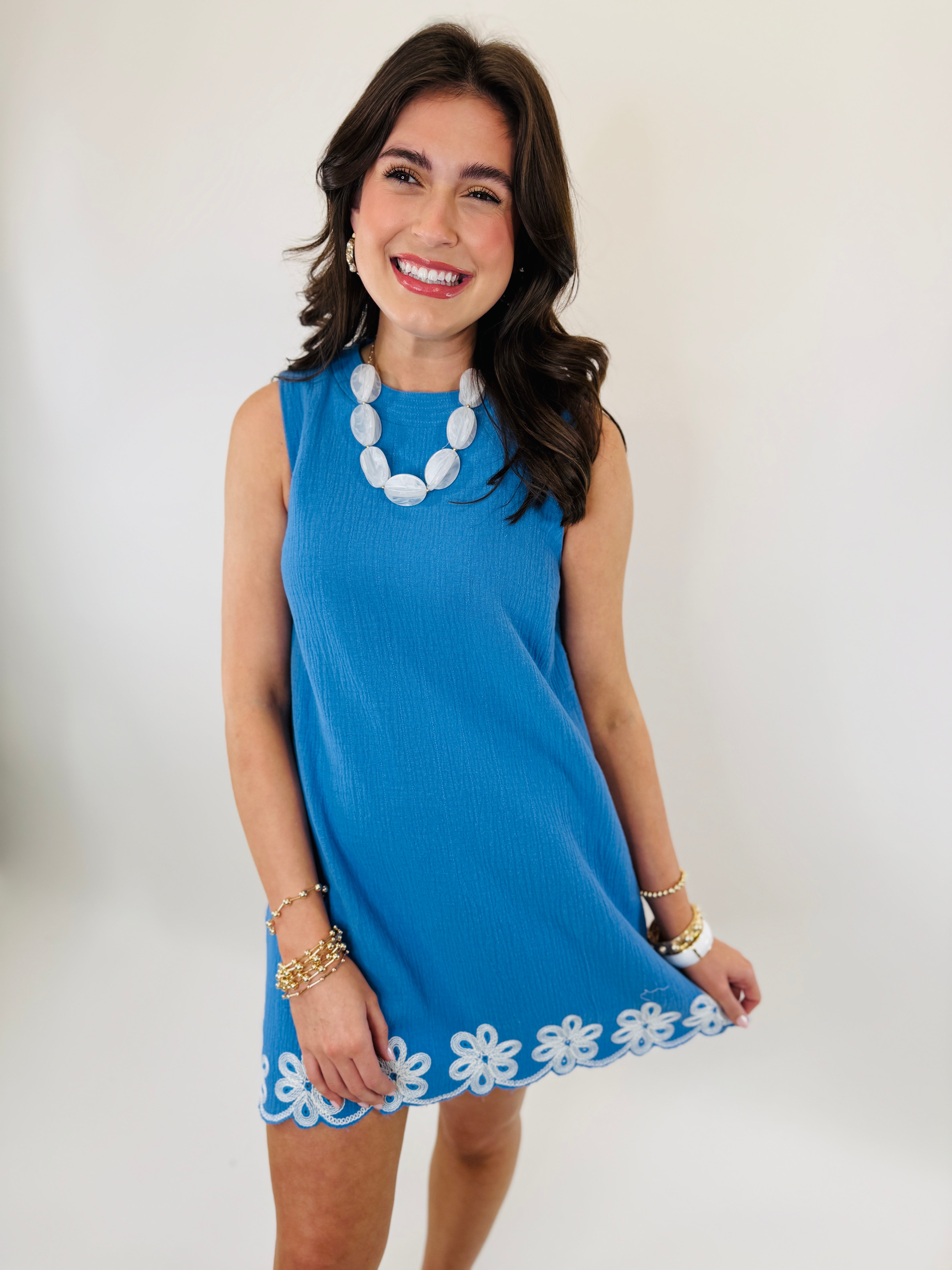 Woman wearing blue sleeveless dress with white floral embroidered hem, white beaded necklace, and gold bracelets.