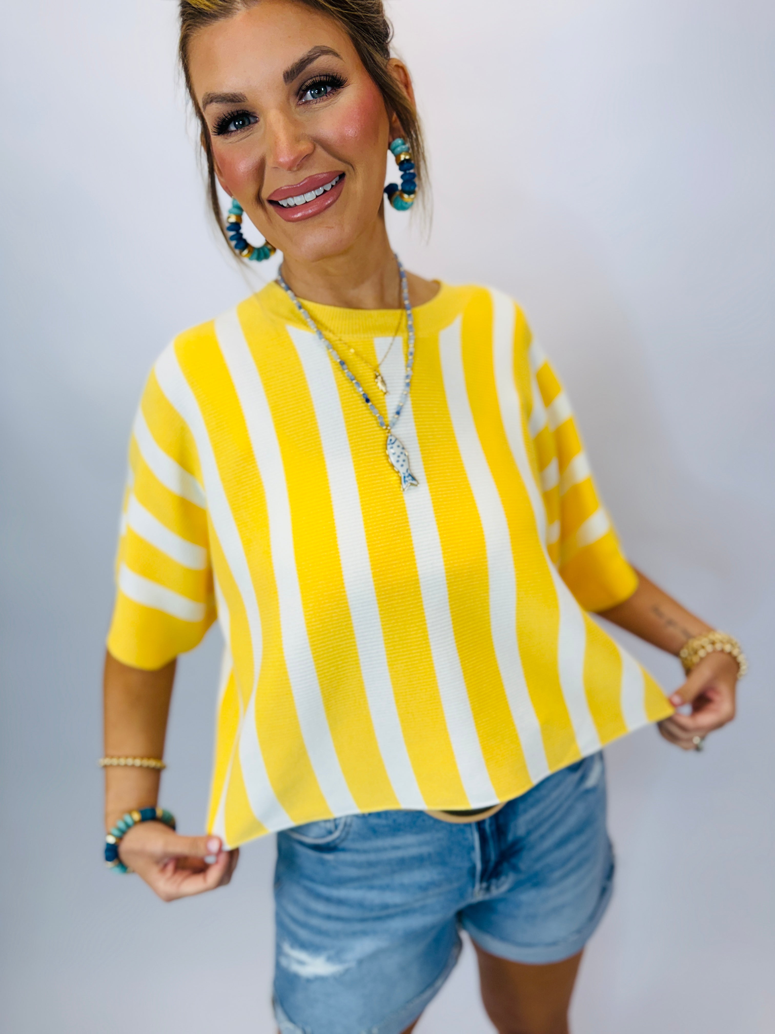 Woman wearing yellow and white striped sweater with three-quarter sleeves, paired with denim shorts and colorful jewelry.
