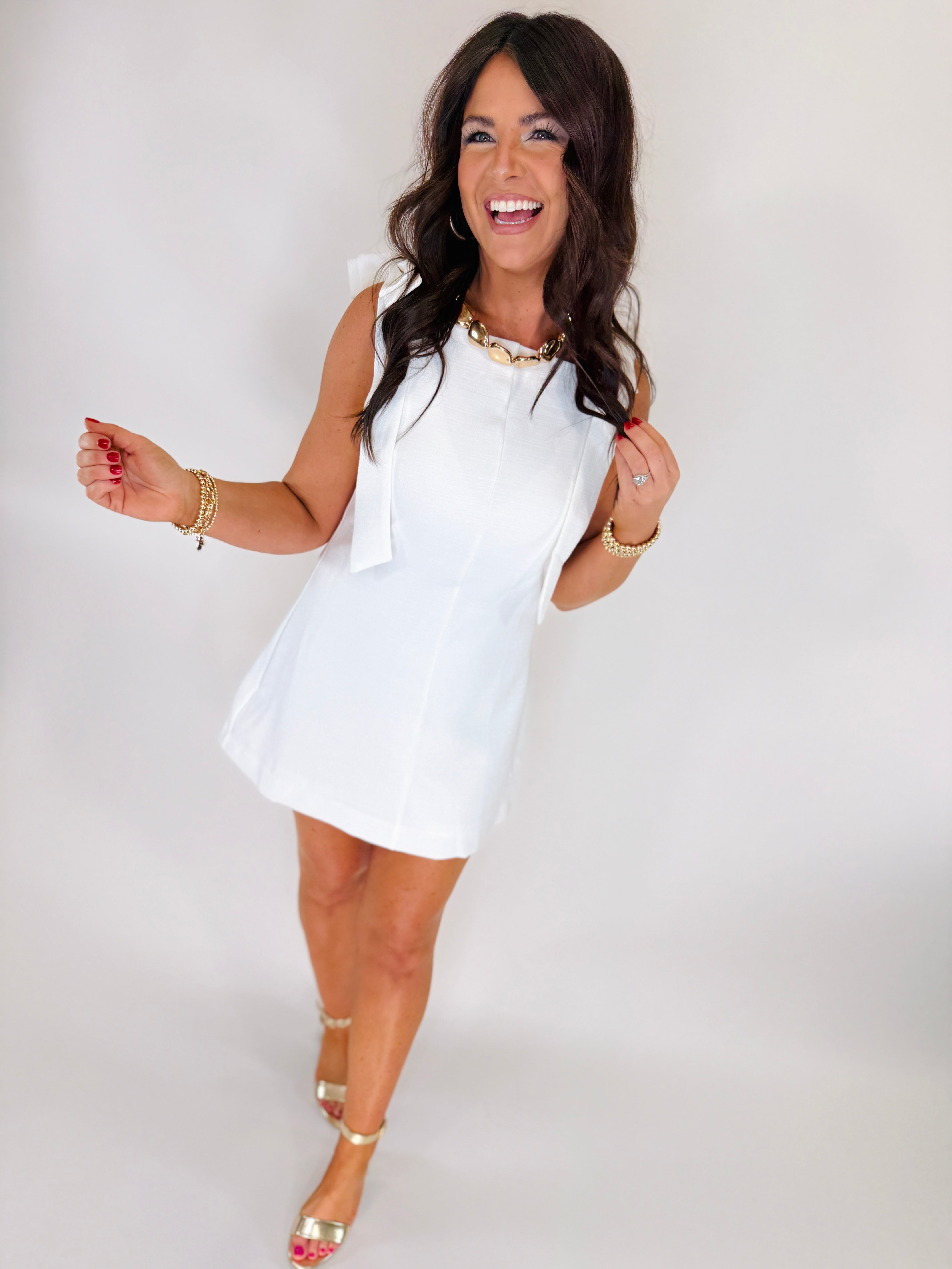 Woman wearing white sleeveless Kennedy Soft Mini Dress with gold jewelry and heels, smiling against white background.