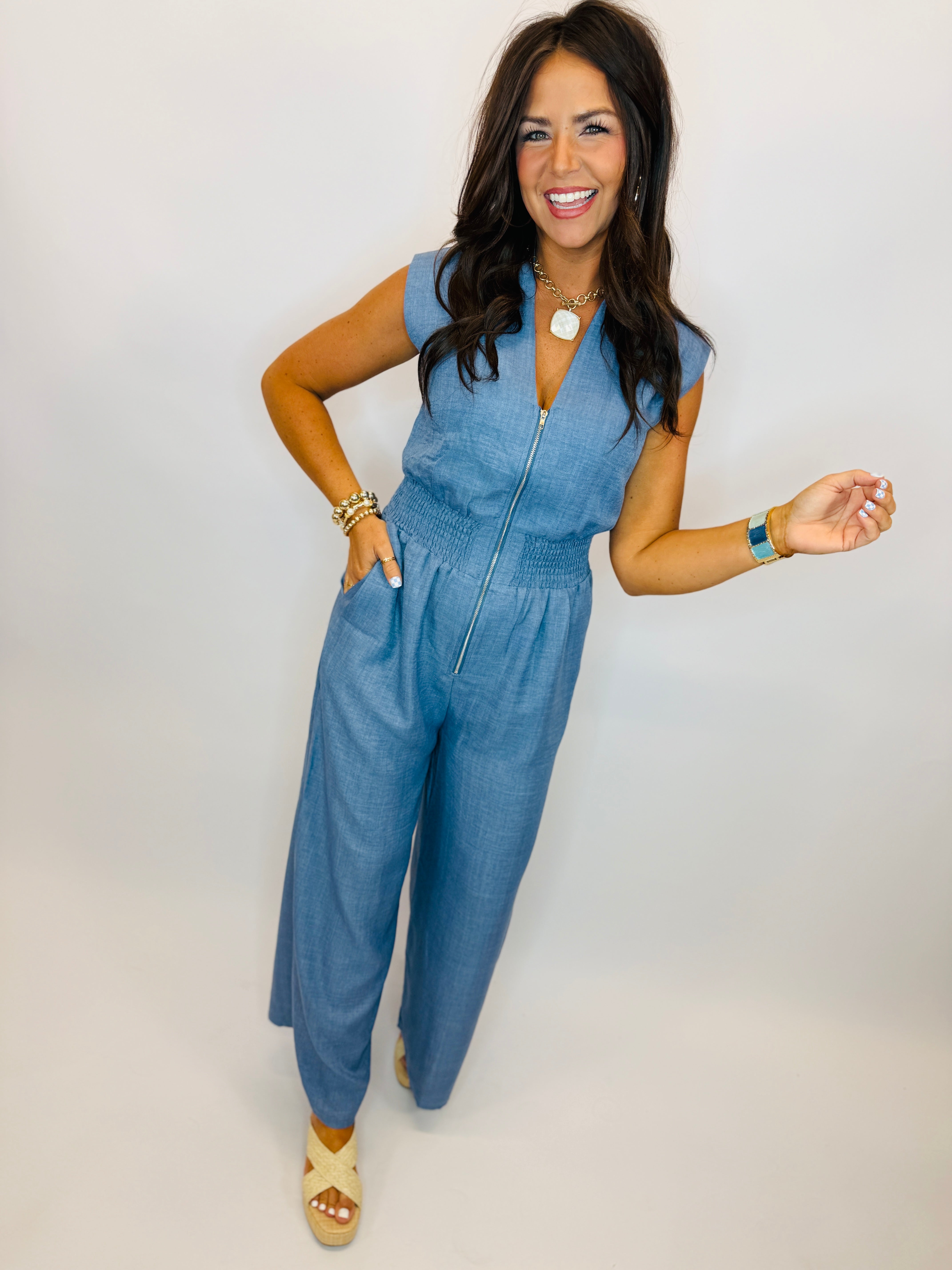 Woman wearing blue denim sleeveless jumpsuit with front zipper and elastic waist, styled with platform sandals