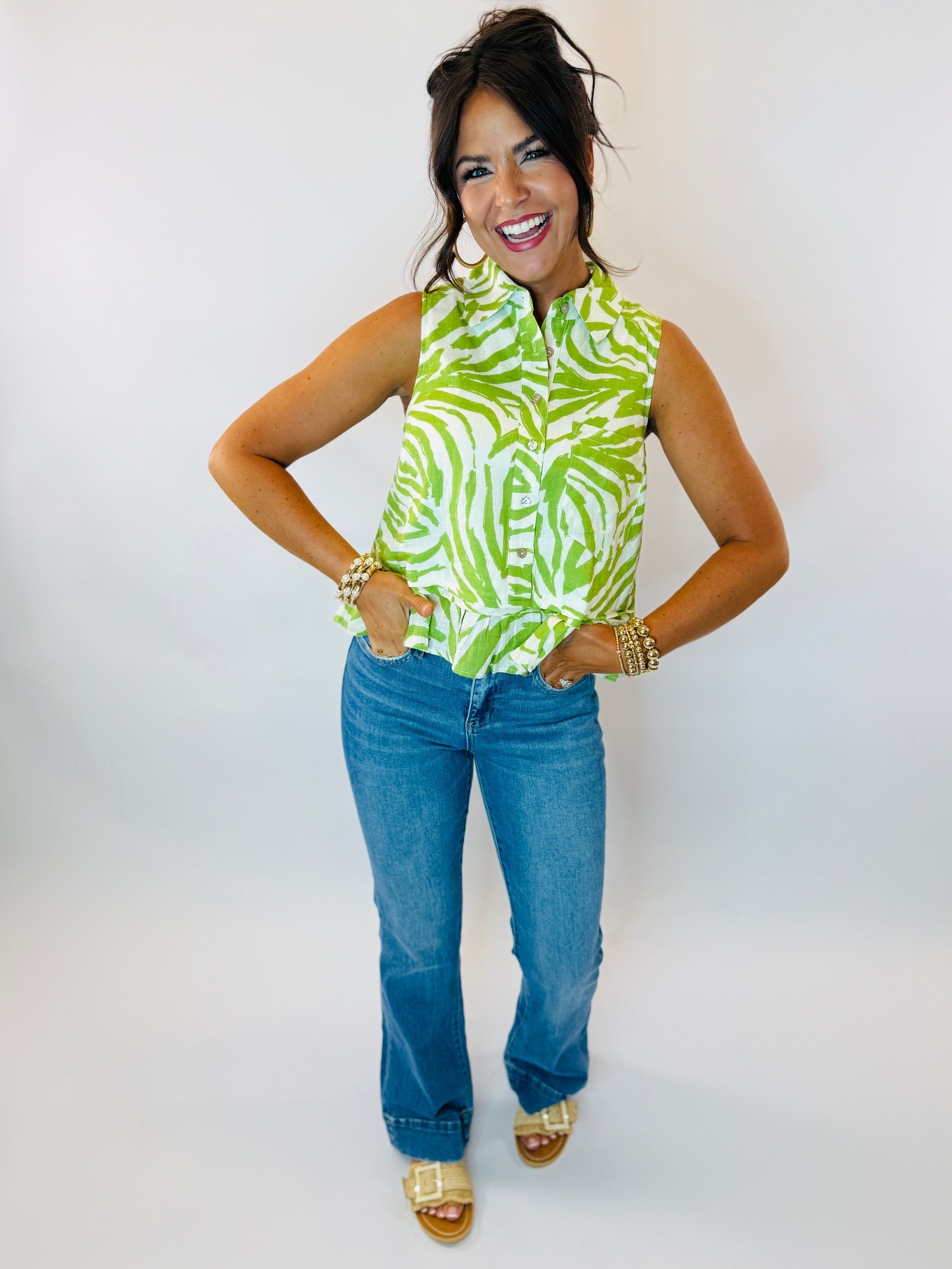 Woman wearing green and white zebra print sleeveless button-up top with blue jeans and gold sandals.