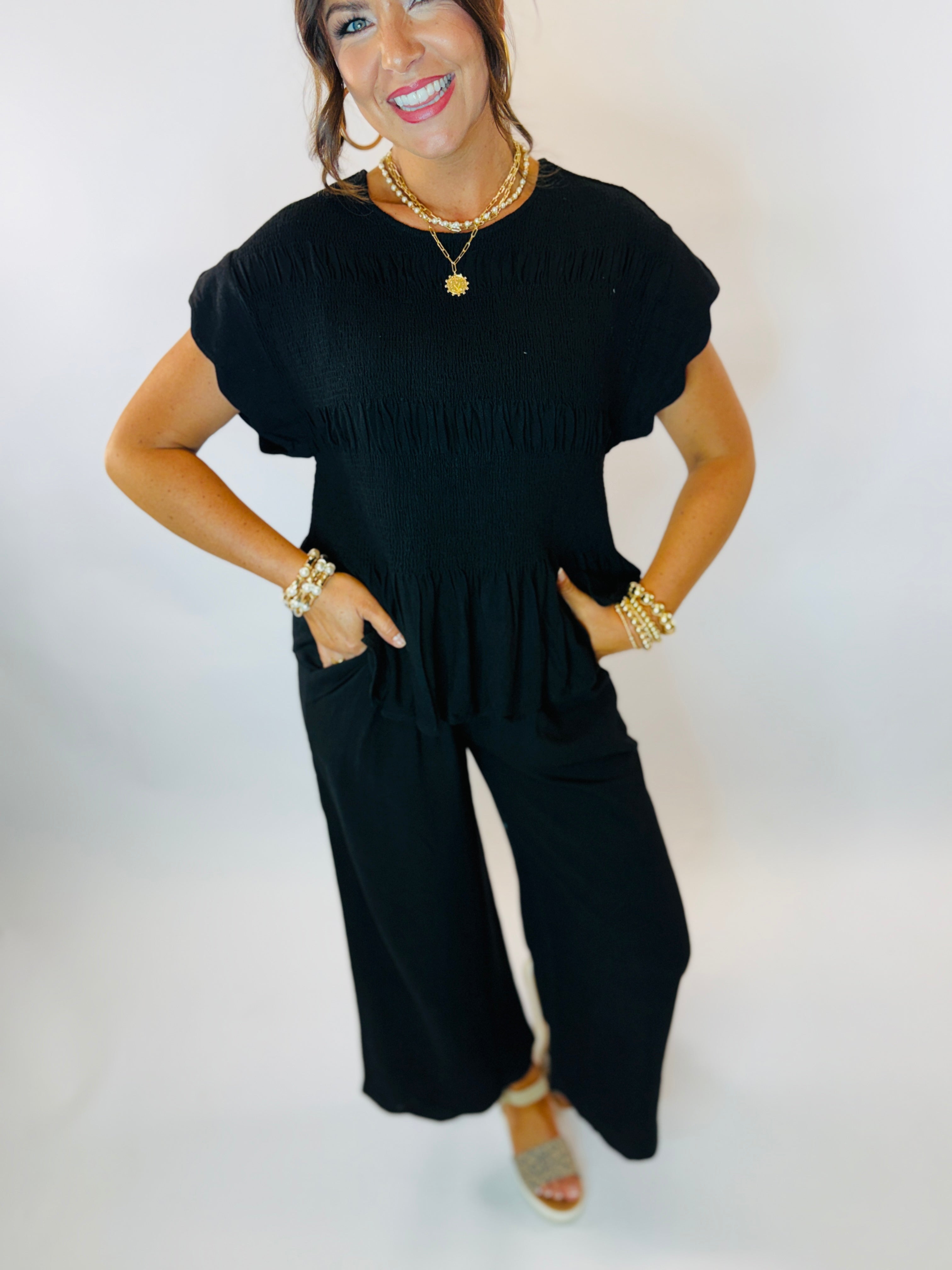 Woman wearing black short-sleeve smocked top with ruffle sleeves, styled with gold jewelry and wide-leg pants.