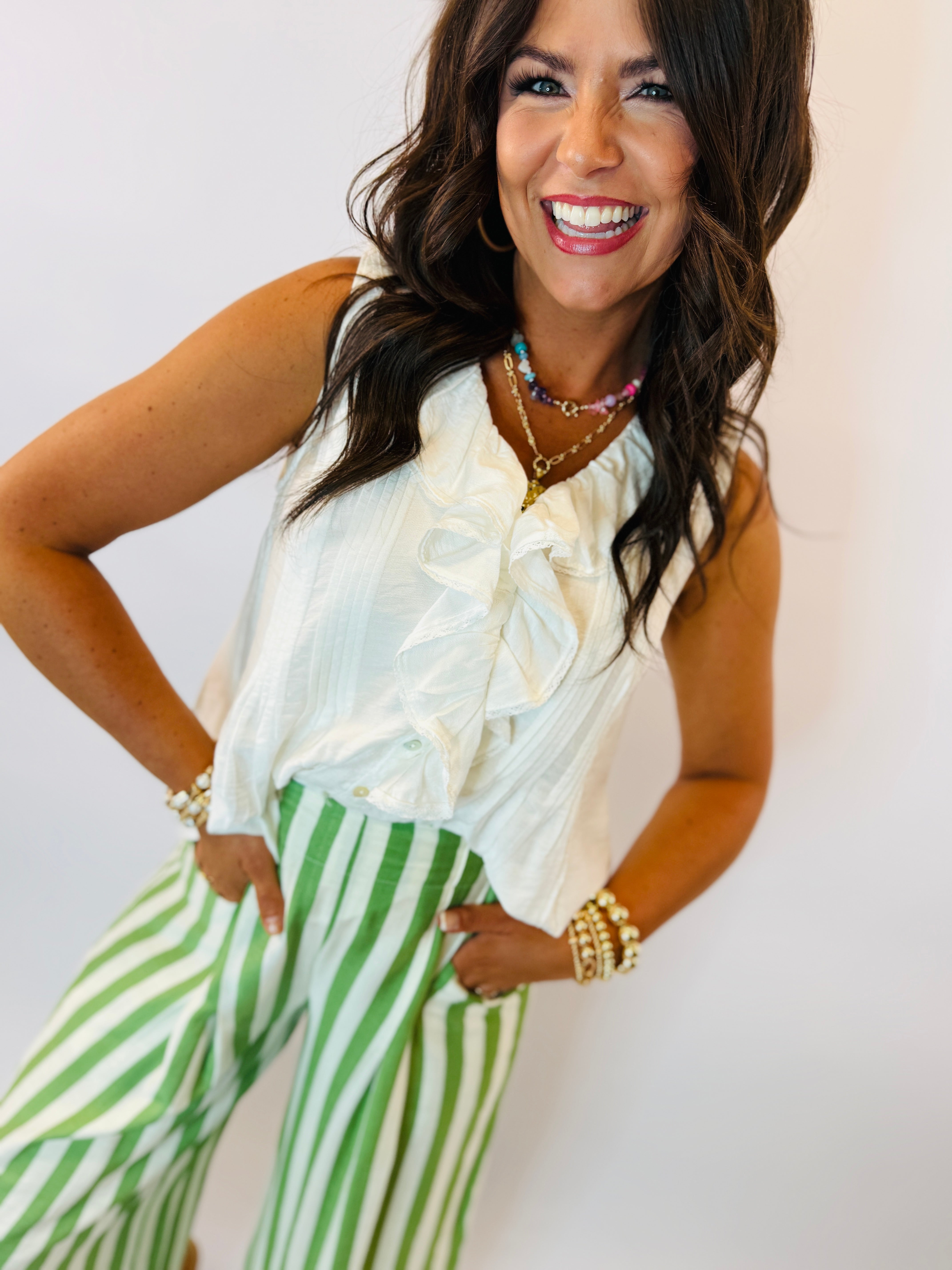 Woman wearing white sleeveless ruffled Ellis top with green and white striped pants, smiling and posing.