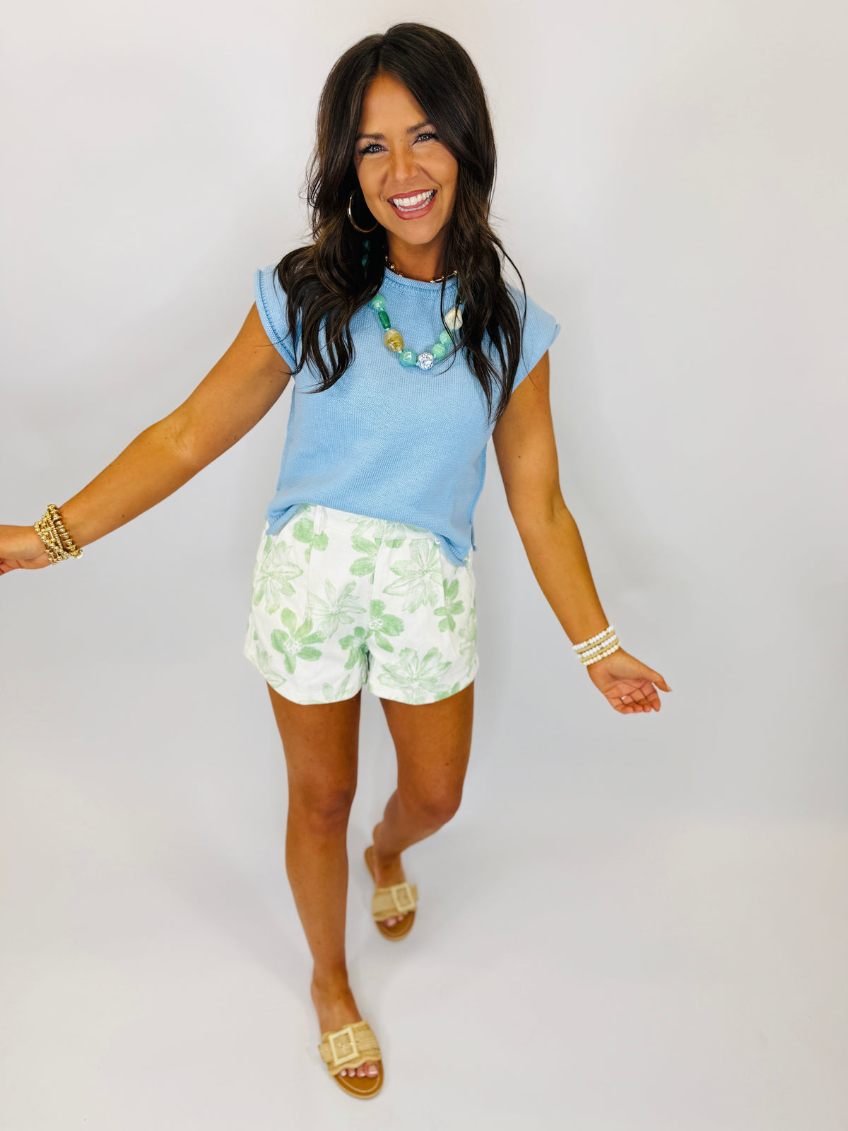 Woman wearing light blue sleeveless top and white shorts with green floral pattern, smiling with arms outstretched.
