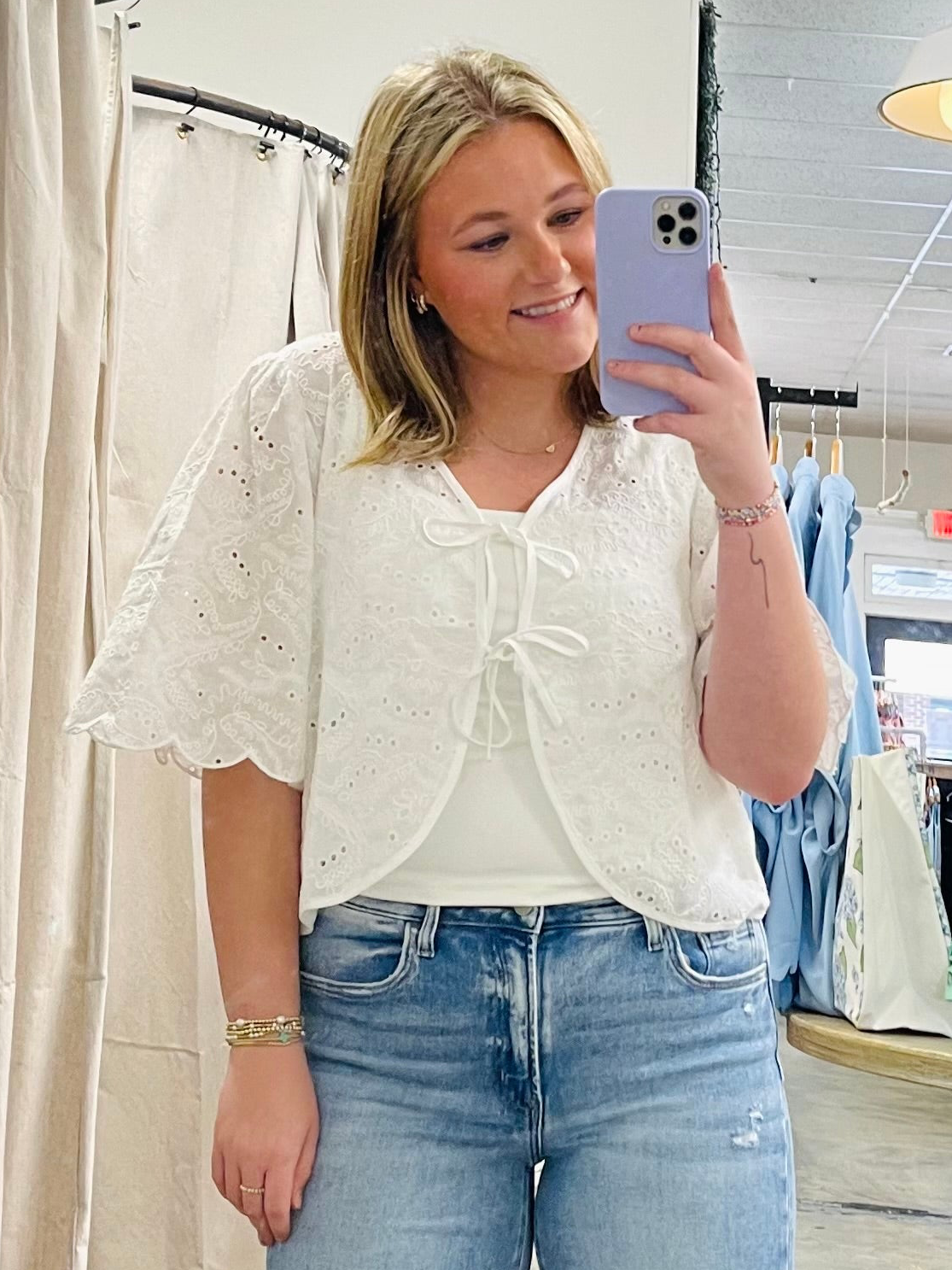 Woman wearing white eyelet lace Hazel top with tie-front detail and short sleeves, paired with blue jeans.