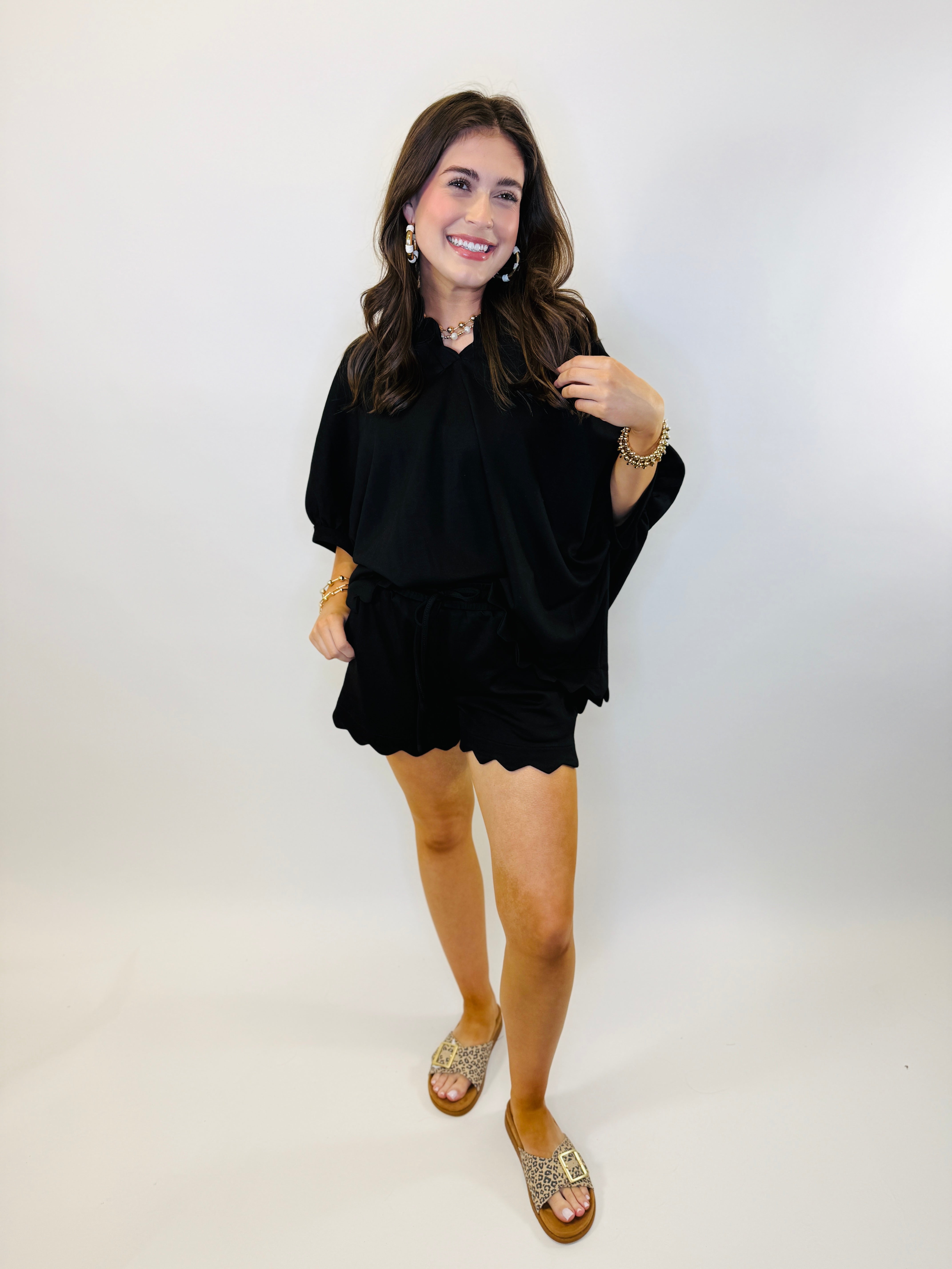 Woman wearing black oversized top with scalloped hem, gold jewelry, and leopard print sandals against white background.