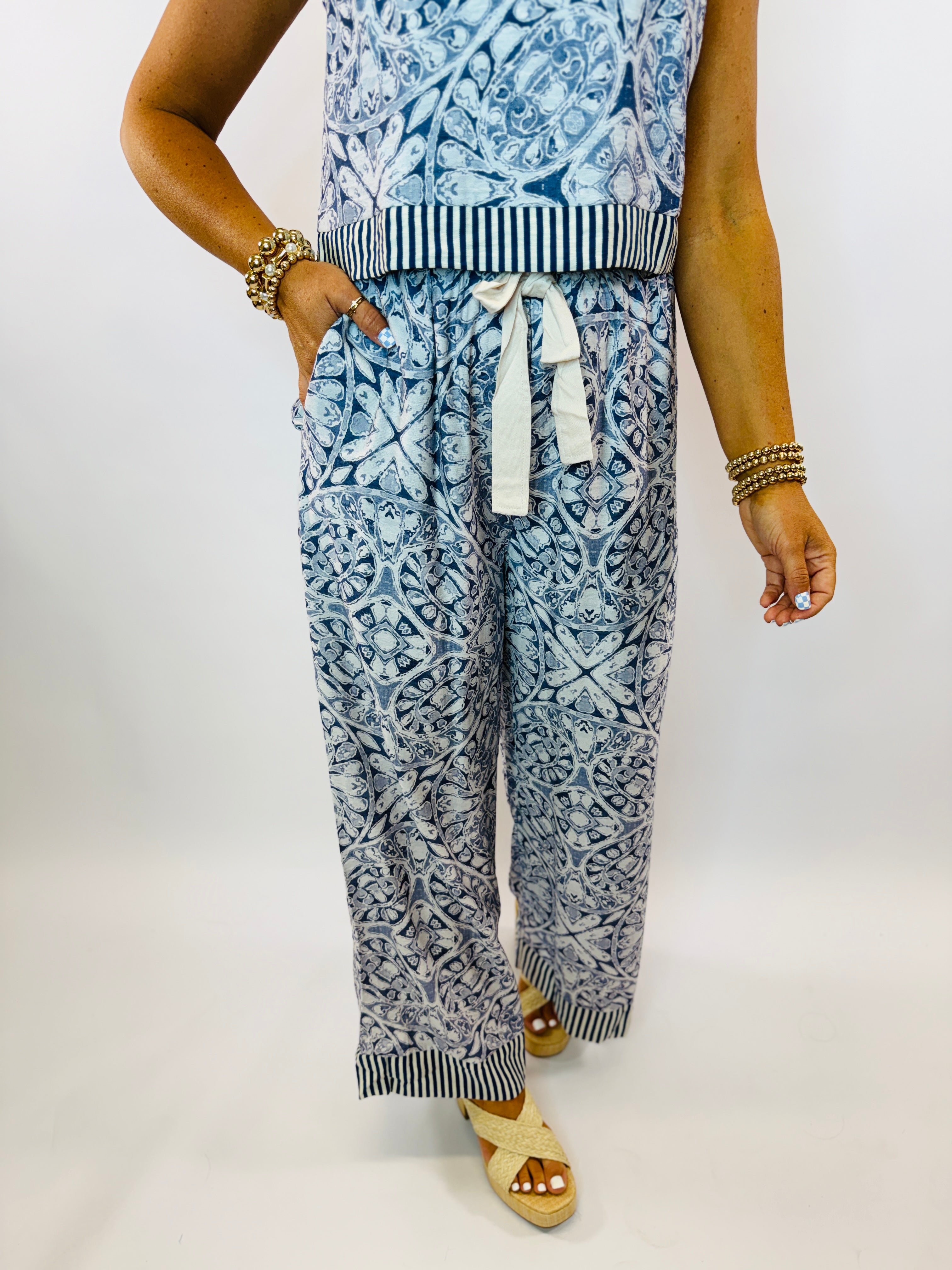 Blue and white patterned wide-leg pants with striped waistband and hem trim, worn with gold jewelry and sandals.