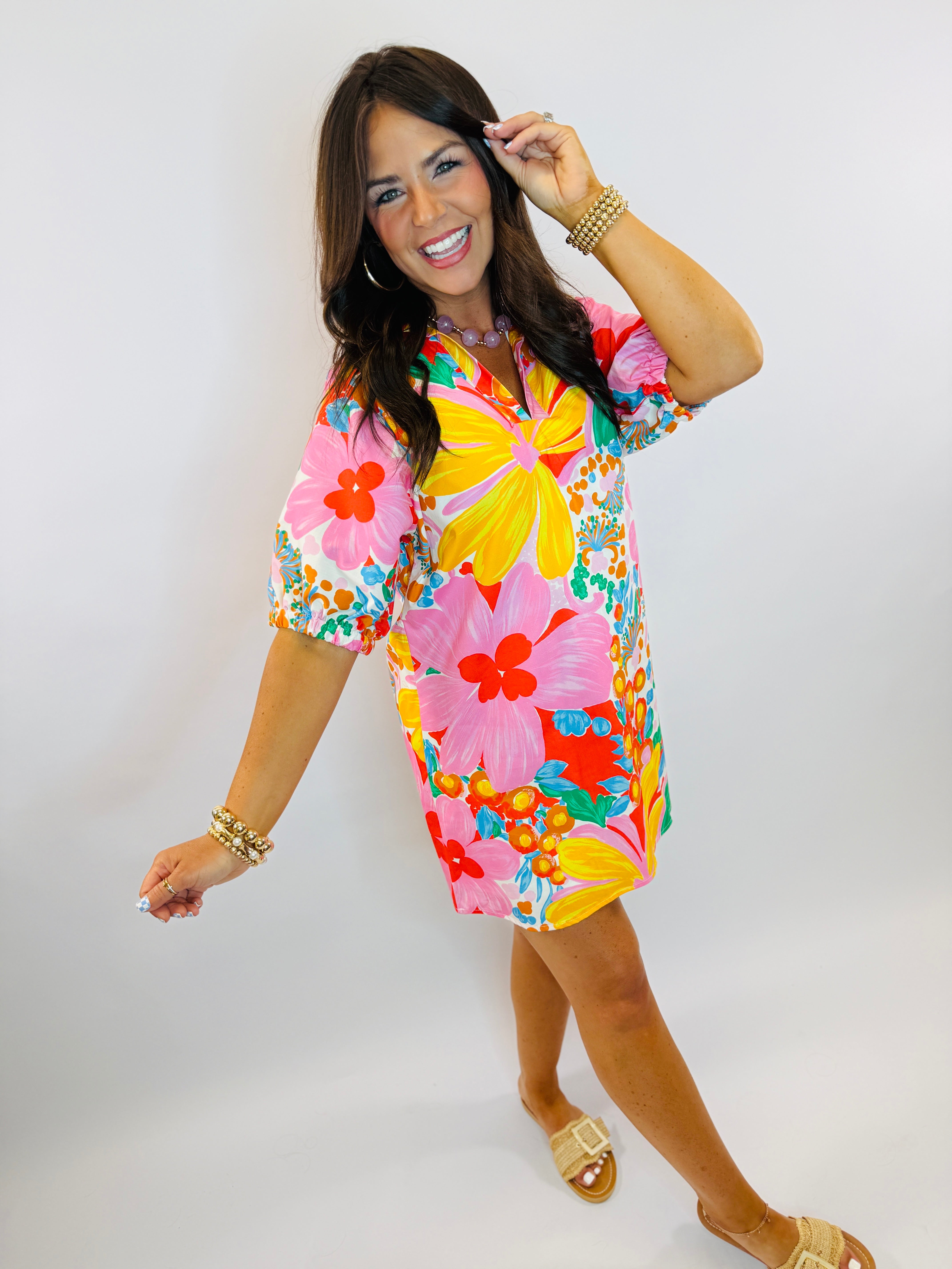 Woman wearing colorful floral print short-sleeve dress with yellow, pink, and red flowers, accessorized with gold jewelry ...