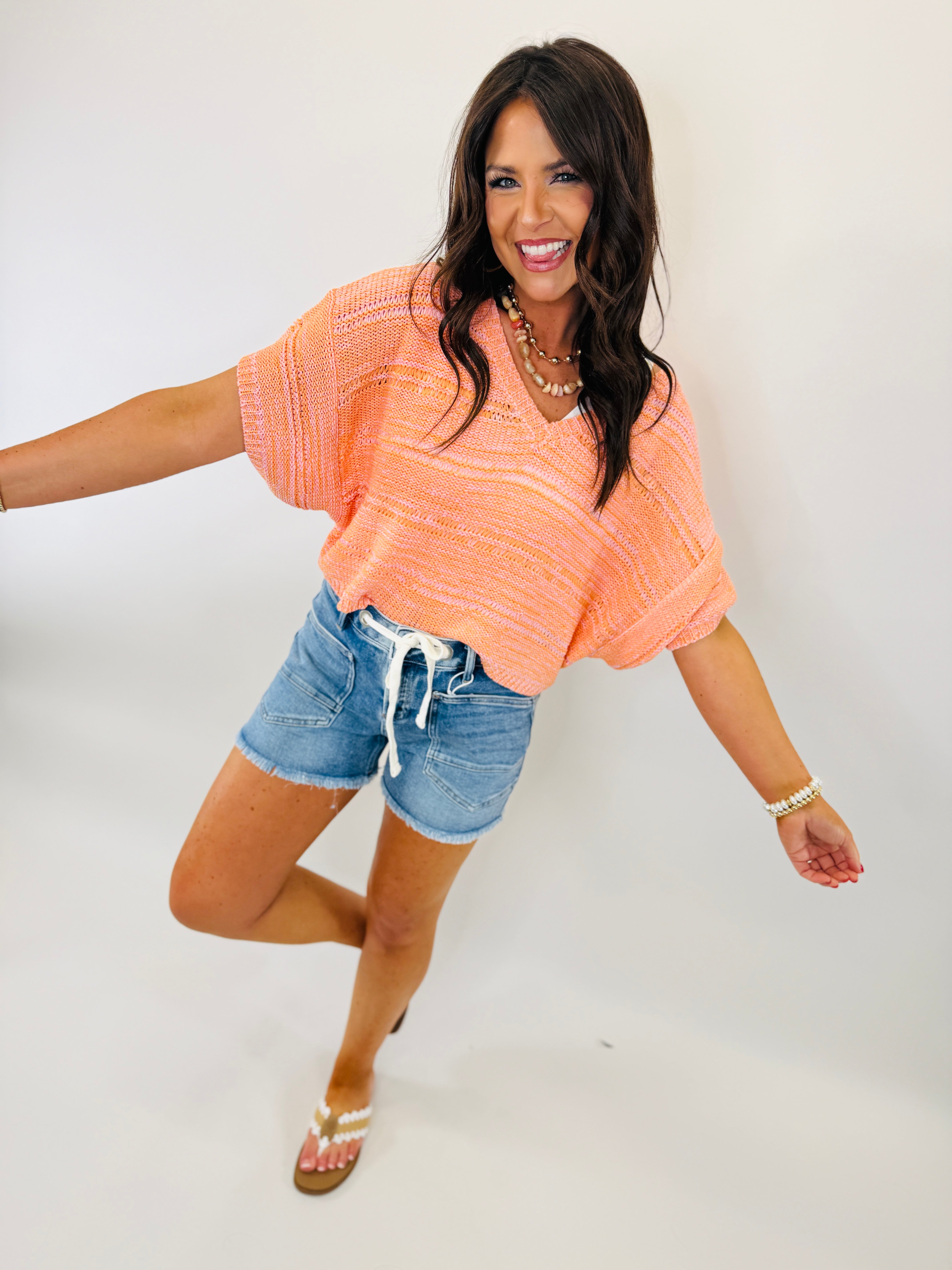Woman wearing coral knit sweater and denim shorts in playful pose with pearl accessories