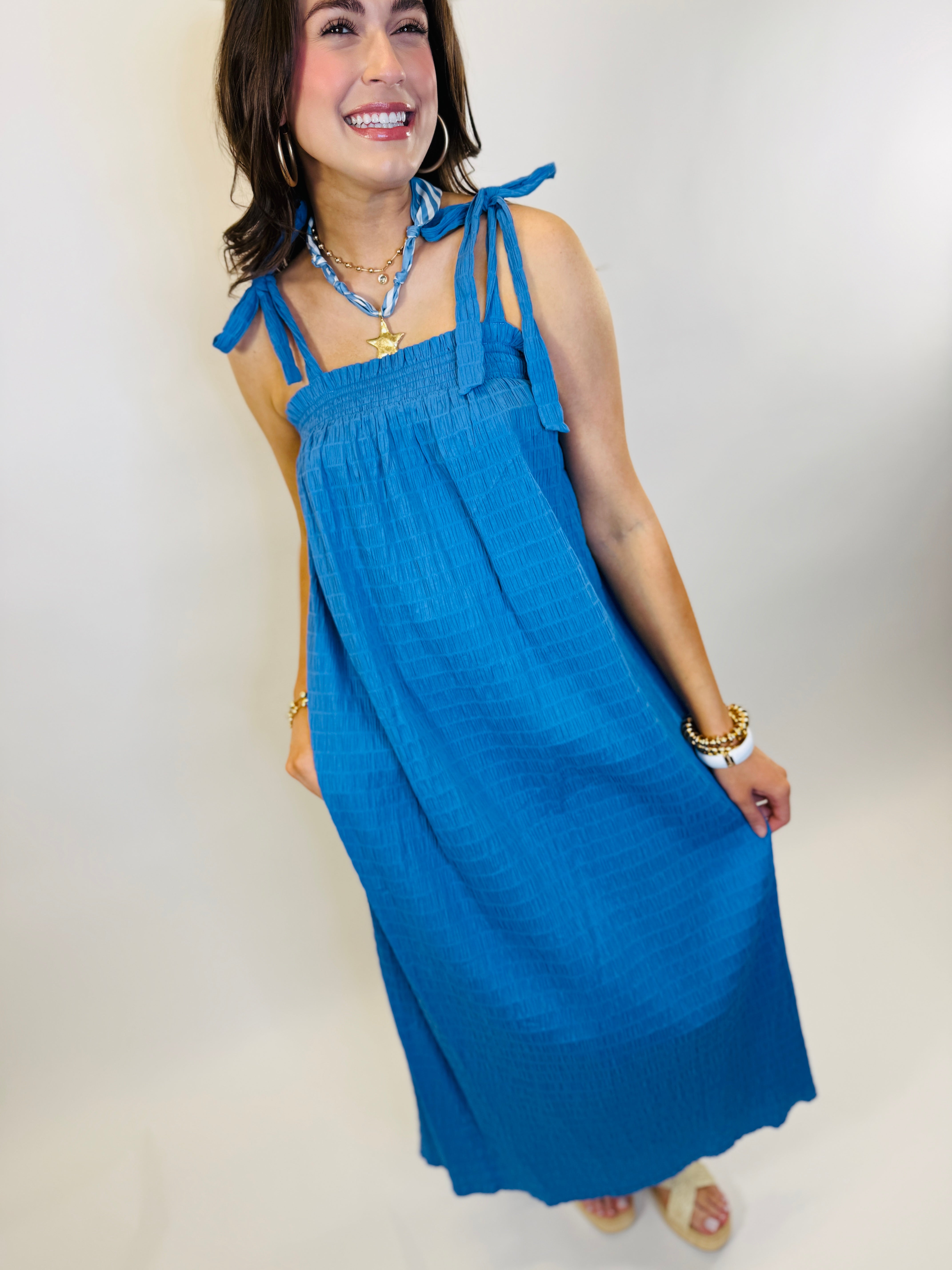 Woman wearing blue halter maxi dress with tie shoulder straps and smocked bodice, styled with gold jewelry.