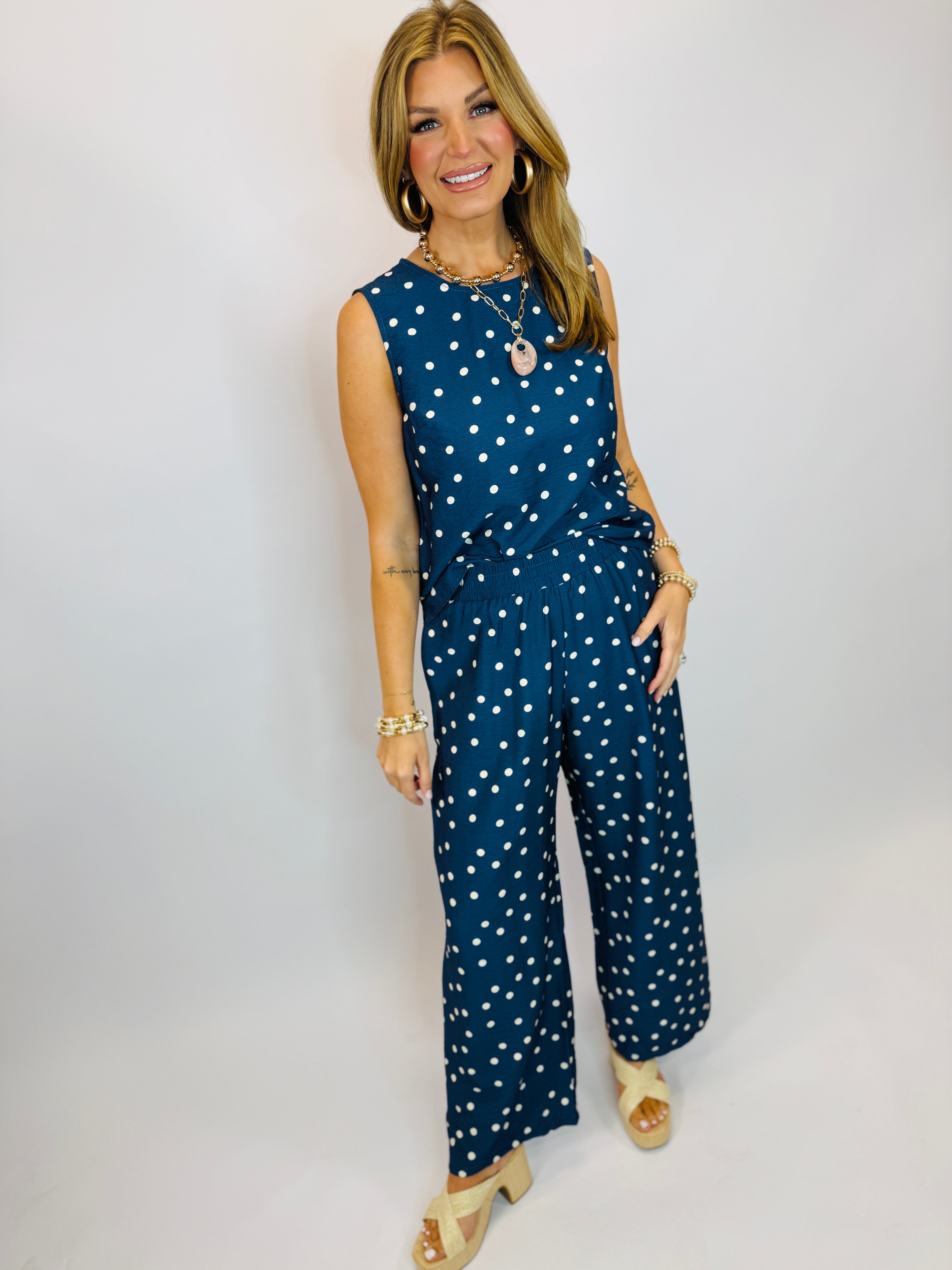 Woman wearing navy blue polka dot sleeveless top and matching wide-leg pants with gold jewelry and sandals.