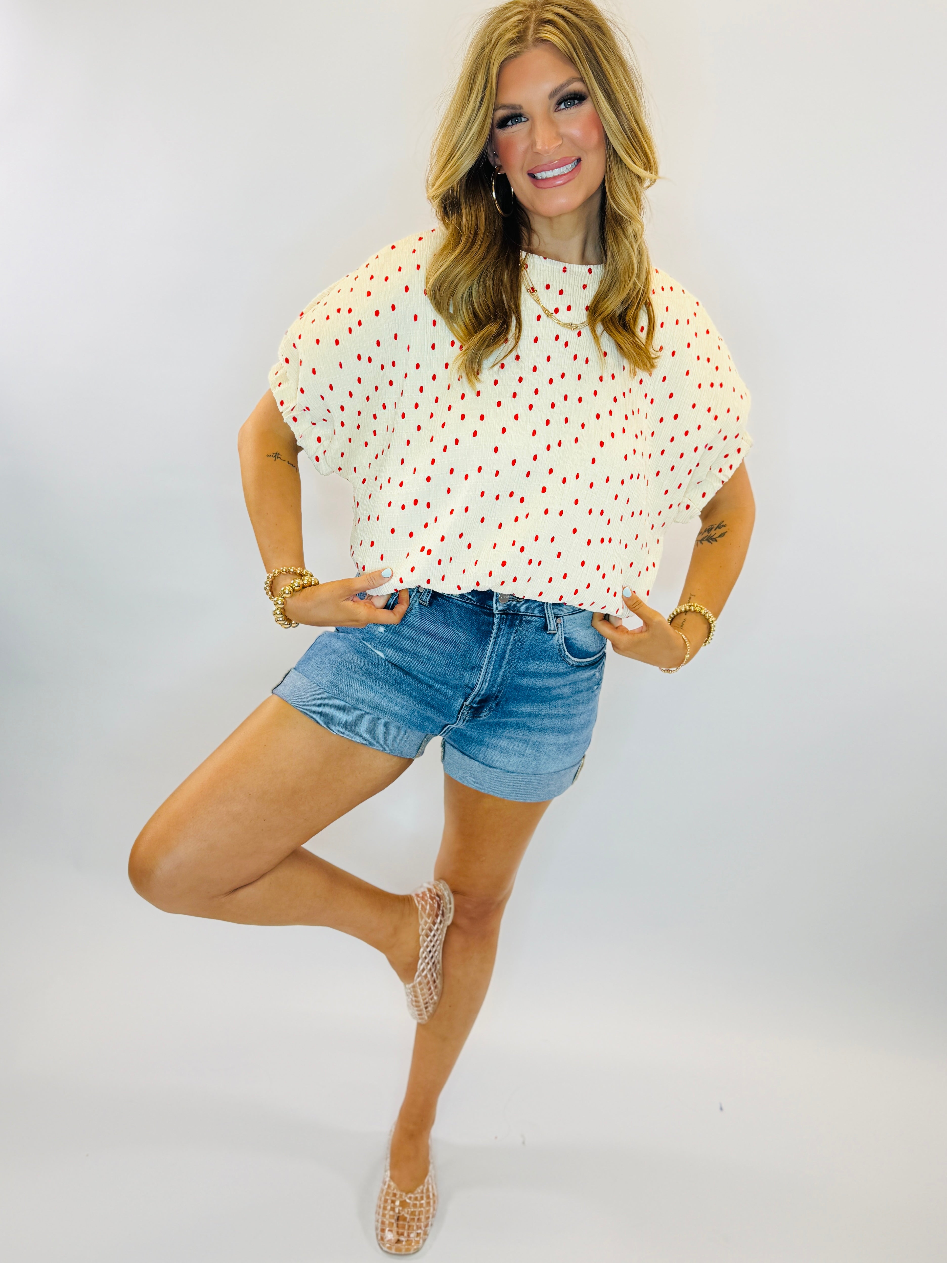 Woman wearing white polka dot short-sleeve top with red dots, paired with denim shorts and beaded sandals.