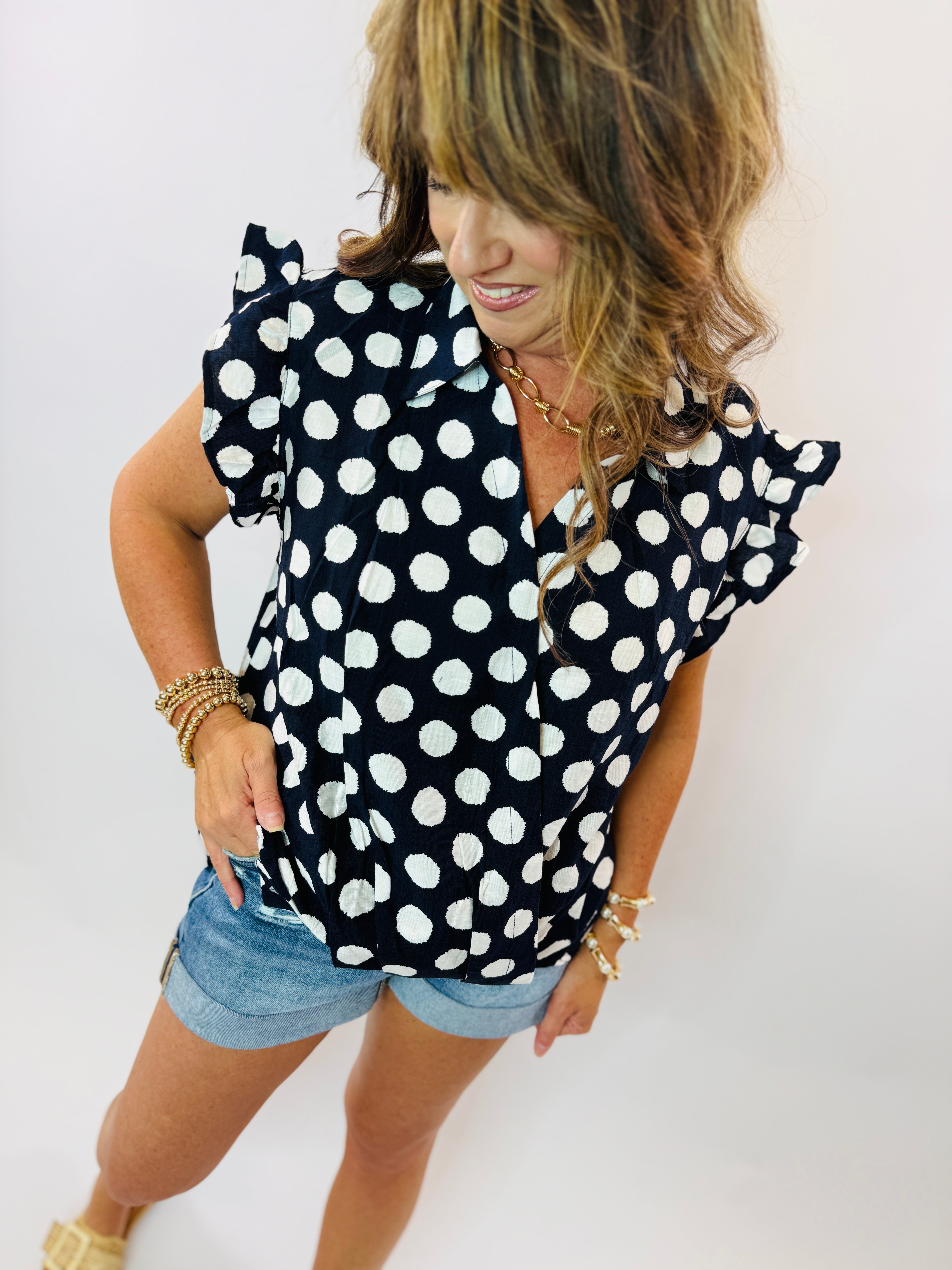 Navy blue polka dot top with ruffle sleeves and V-neckline, styled with denim shorts and gold jewelry.
