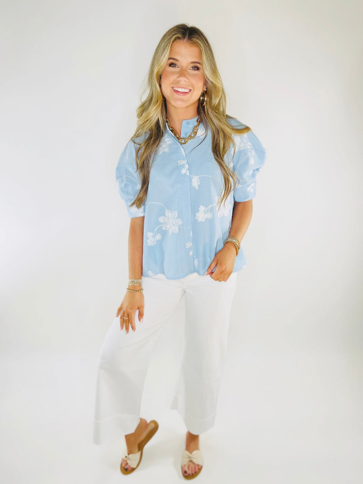 Woman wearing light blue floral button-up shirt with puffed sleeves, white pants, and gold jewelry accessories