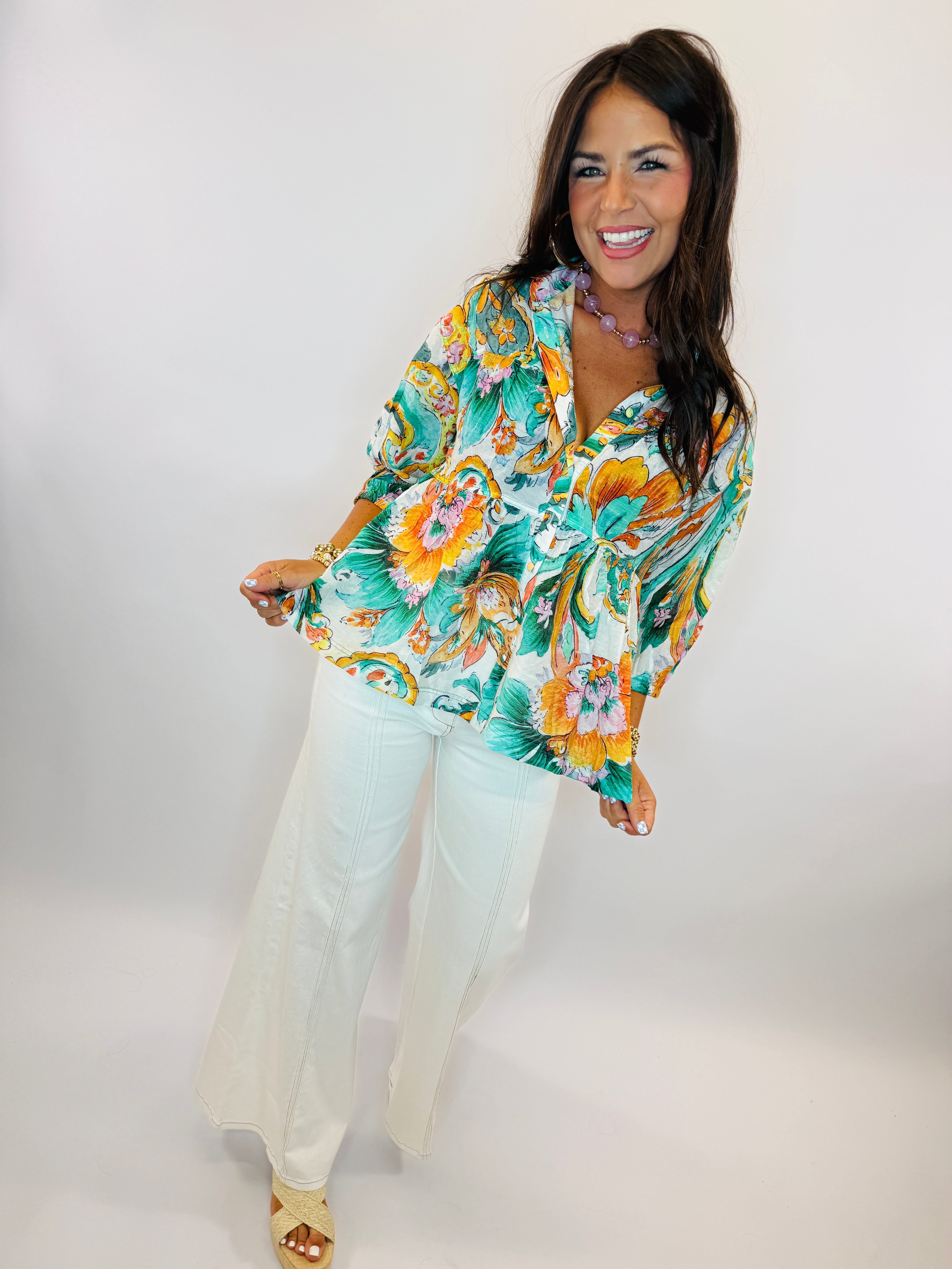 Woman wearing vibrant teal floral print long-sleeve blouse with white jeans, smiling against neutral background.
