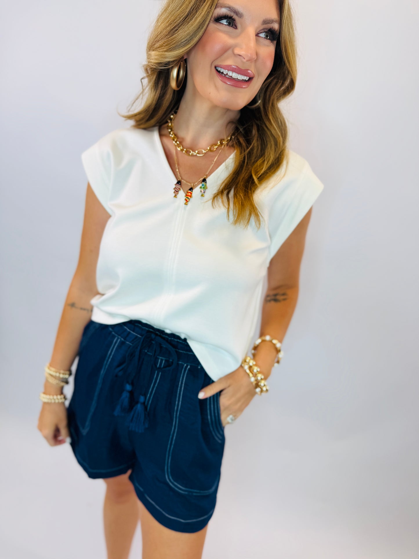 Woman wearing white cap-sleeve Meridian top with gold chain necklace and navy shorts, smiling against white background.