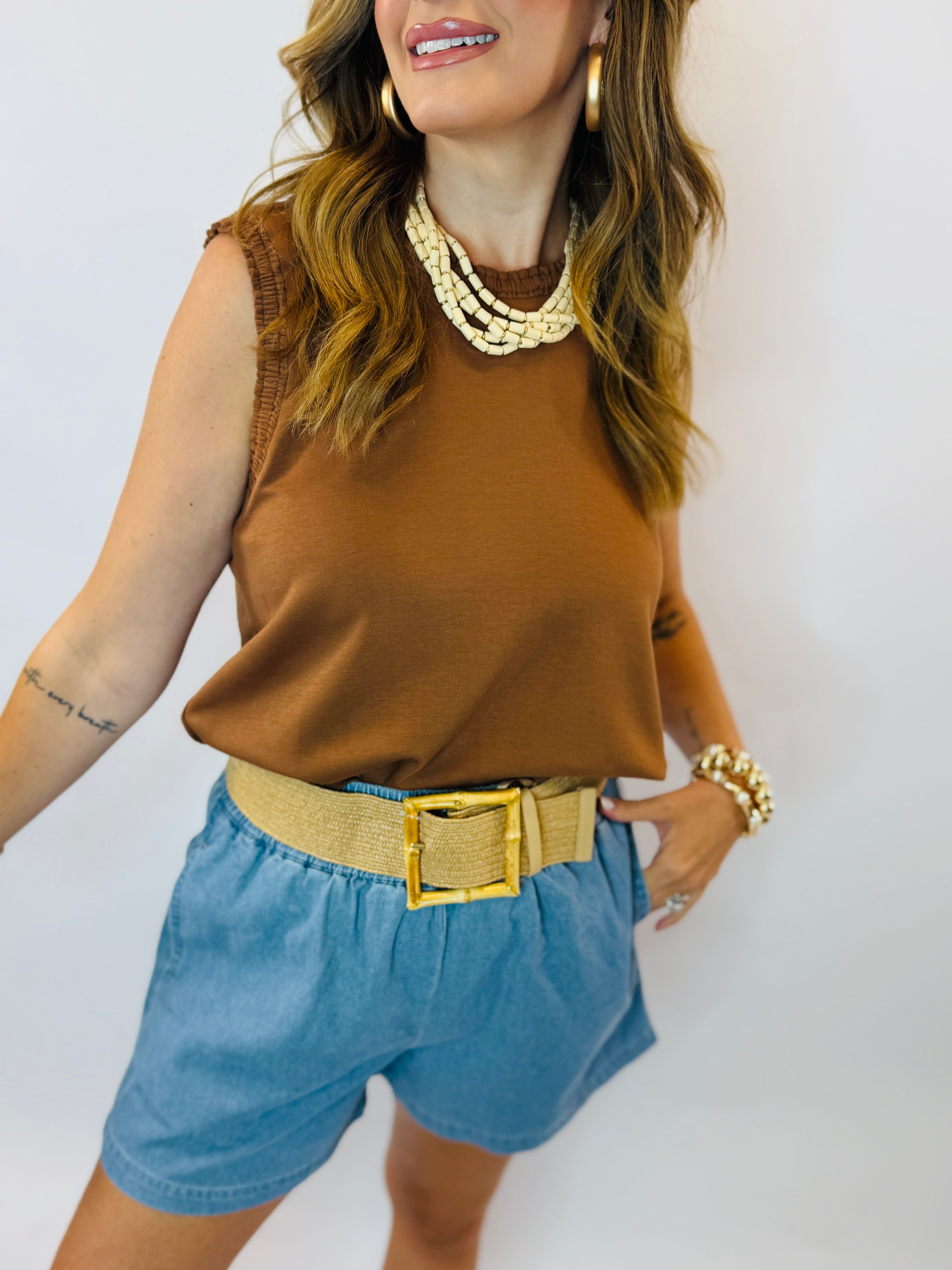 Woman wearing brown sleeveless top and blue denim Joni pull-on shorts with gold belt and pearl necklace.
