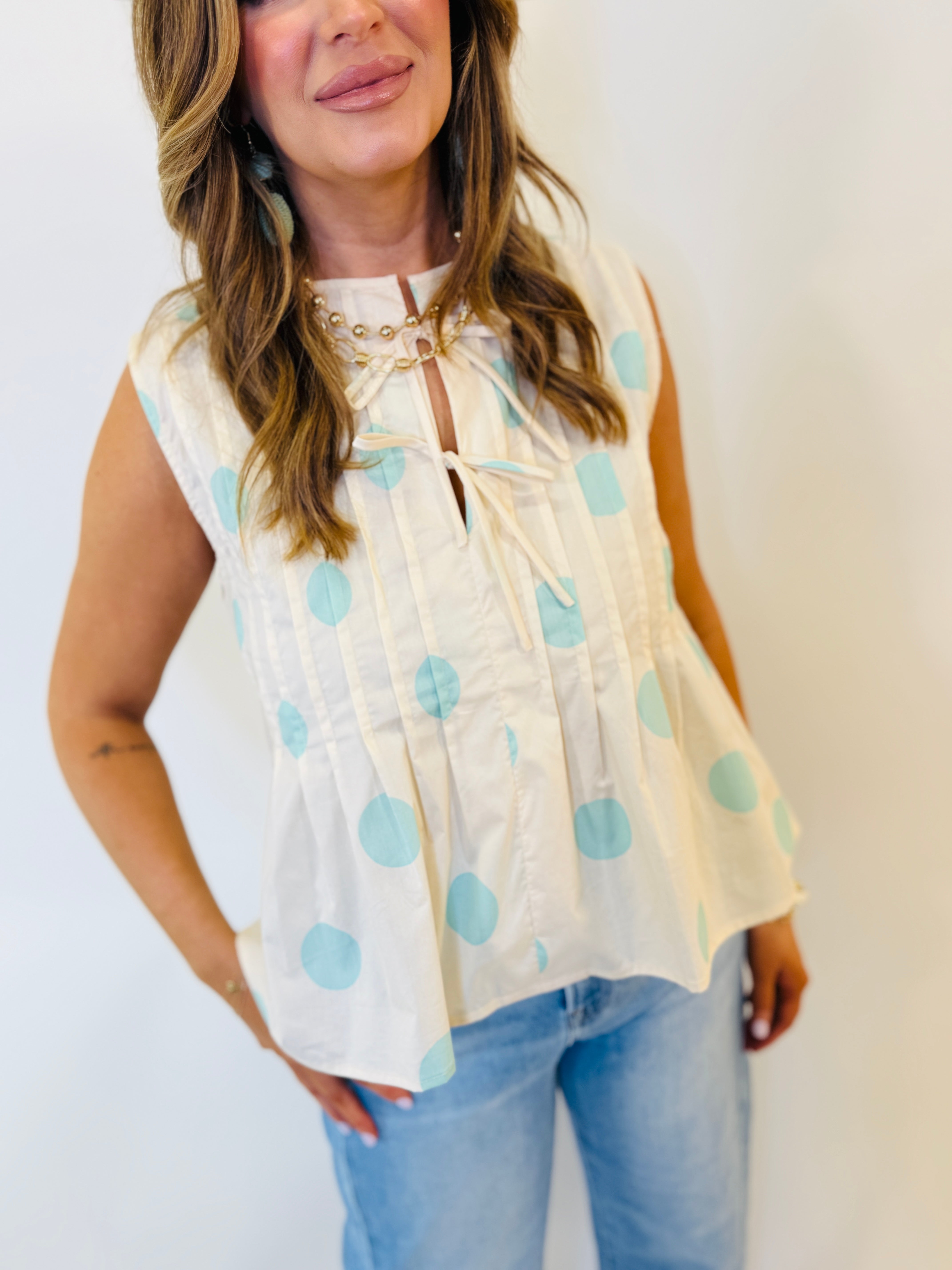 White sleeveless blouse with light blue polka dots, keyhole neckline, and gold chain detail worn with jeans.