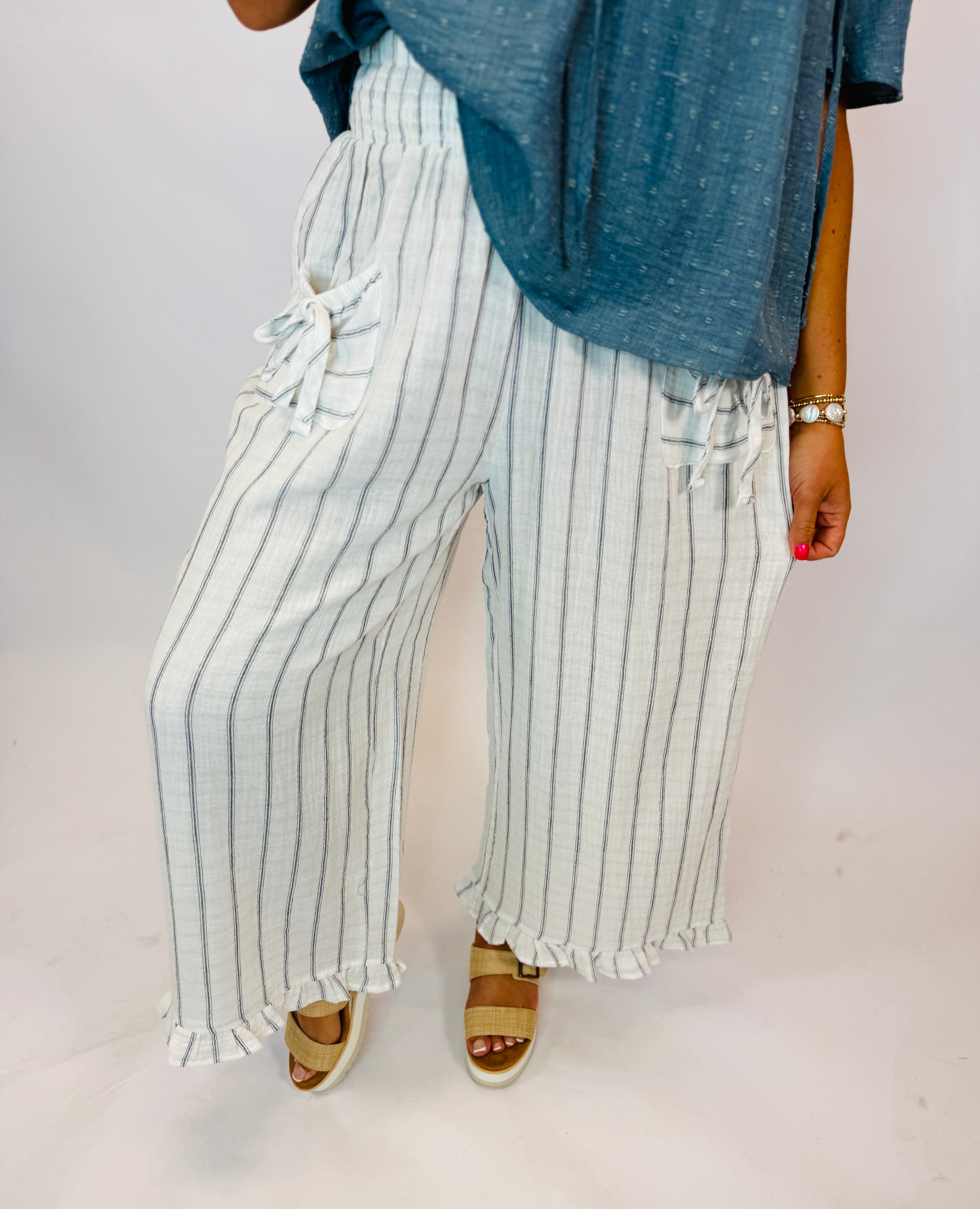 Wide-leg white linen pants with navy pinstripes, ruffle hem, drawstring waist, styled with blue top and sandals.