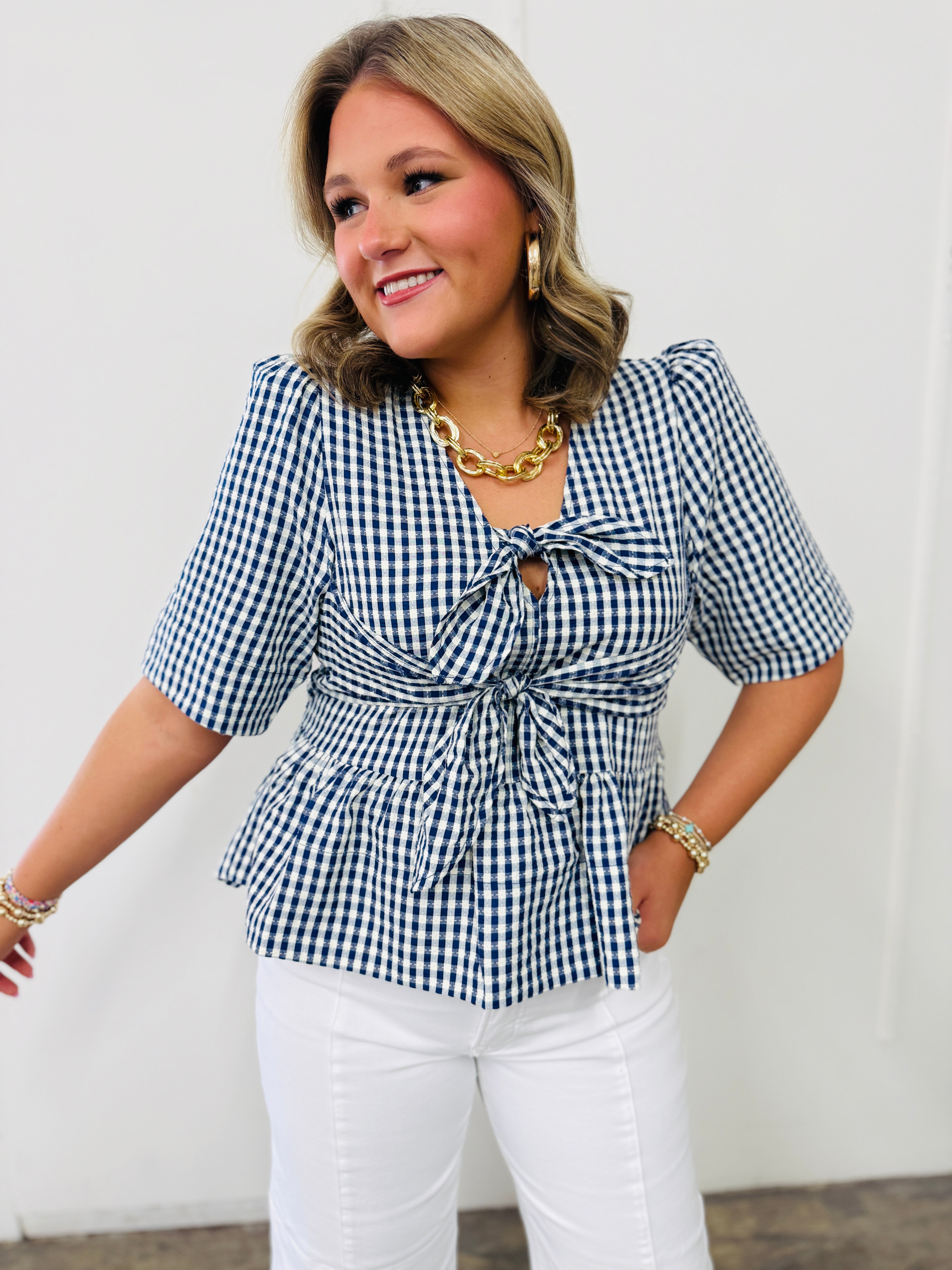 Woman wearing blue and white gingham checked flowy top with tie front detail and white pants