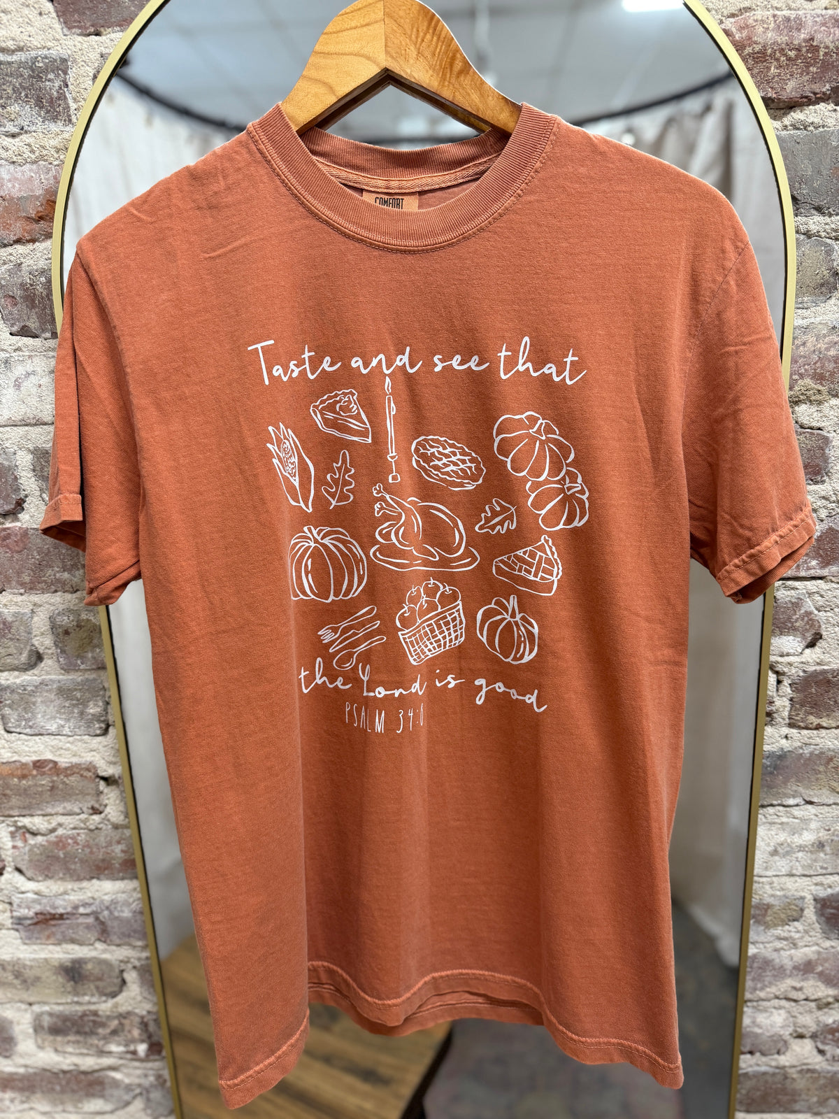 Rust orange t-shirt with "Taste and see that the Lord is good" text and fall food illustrations hanging on wooden hanger